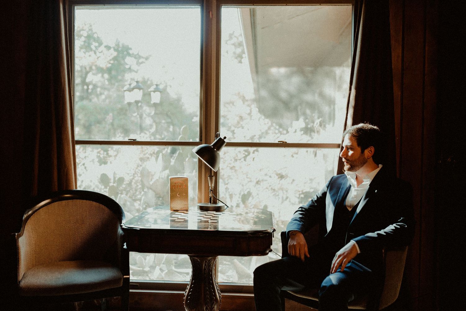 A groom in a dark suit sits contemplatively by a large window at a small table with a lamp.