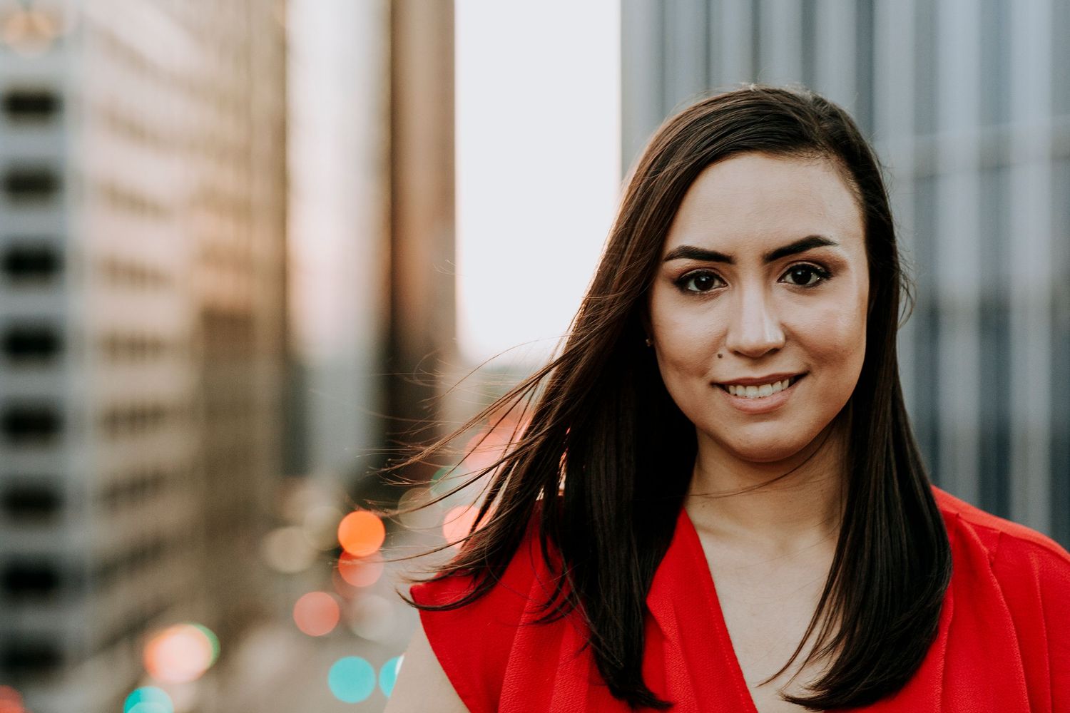 Smiling young woman with dark hair wearing a red jacket, posing outdoors in an urban city setting.