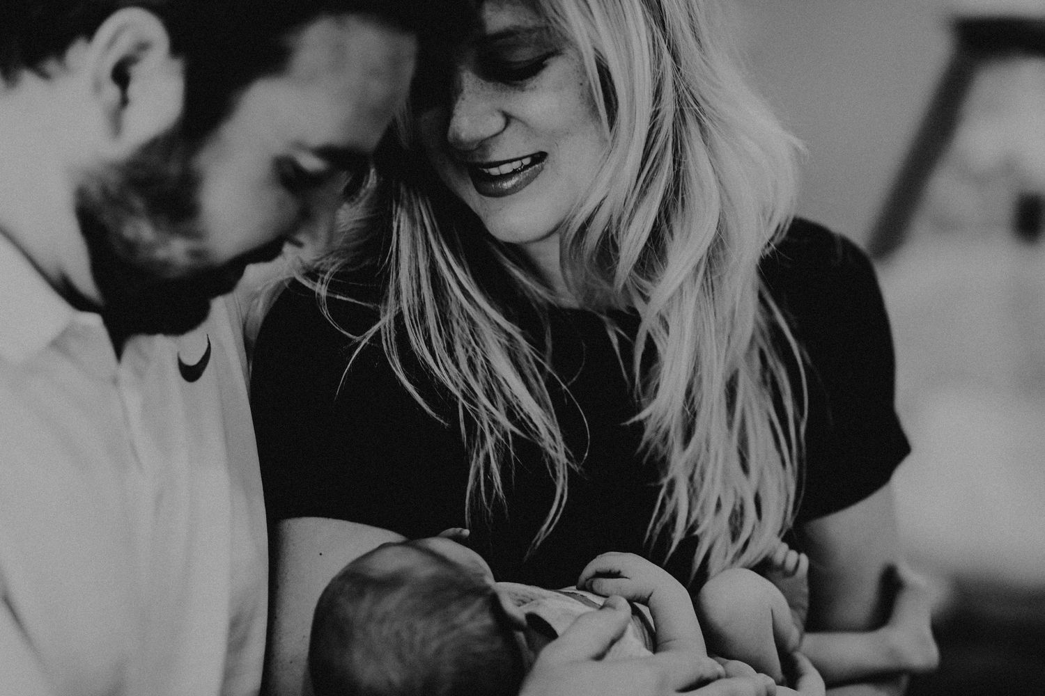 Black and white photo of smiling parents lovingly gazing at their newborn baby.