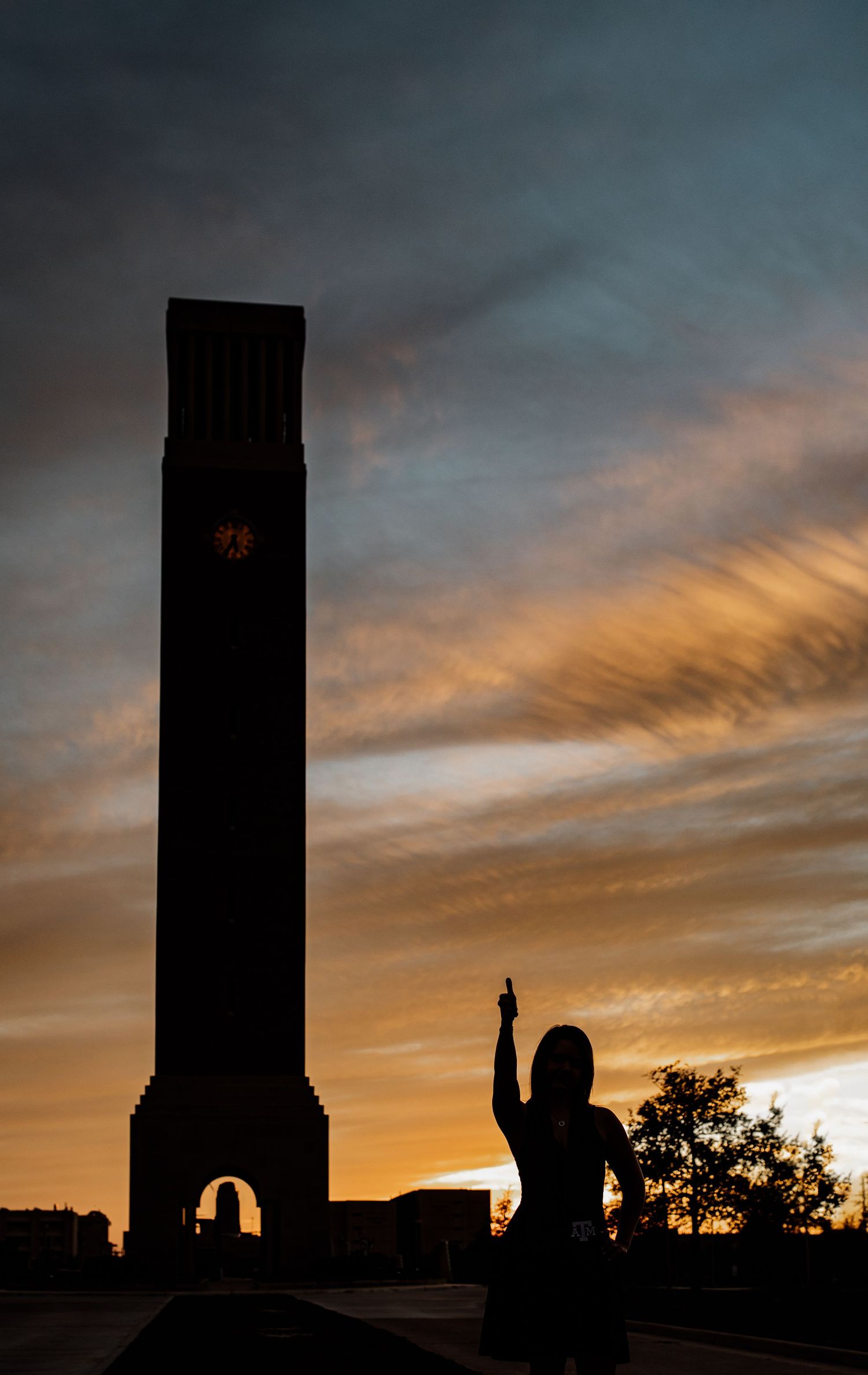 Silhouette of a woman raising her finger beside a tall clock tower against a dramatic orange and blue sunset sky.