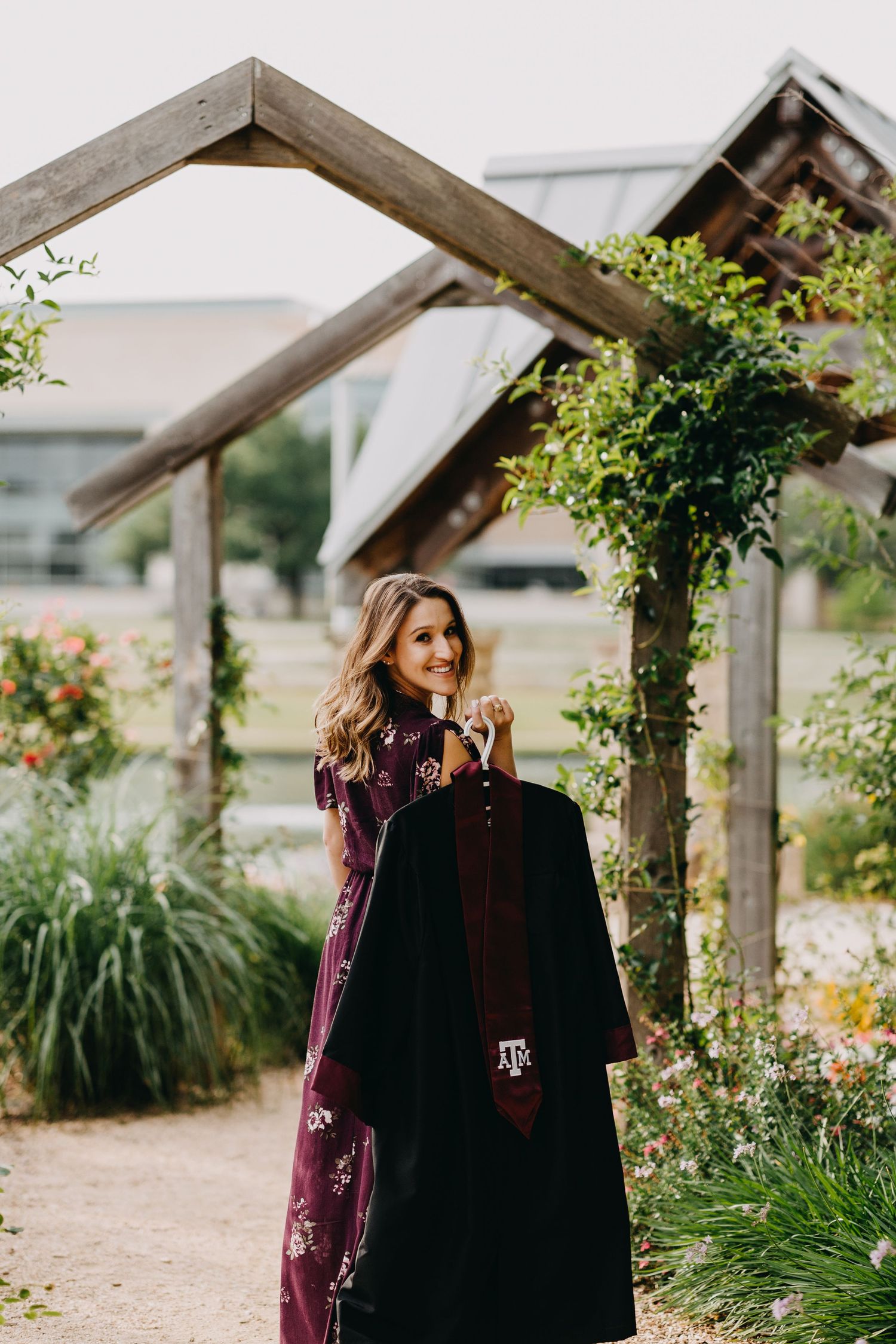 A smiling woman holds a Texas A&M graduation gown in a garden with wooden arbors and lush greenery.