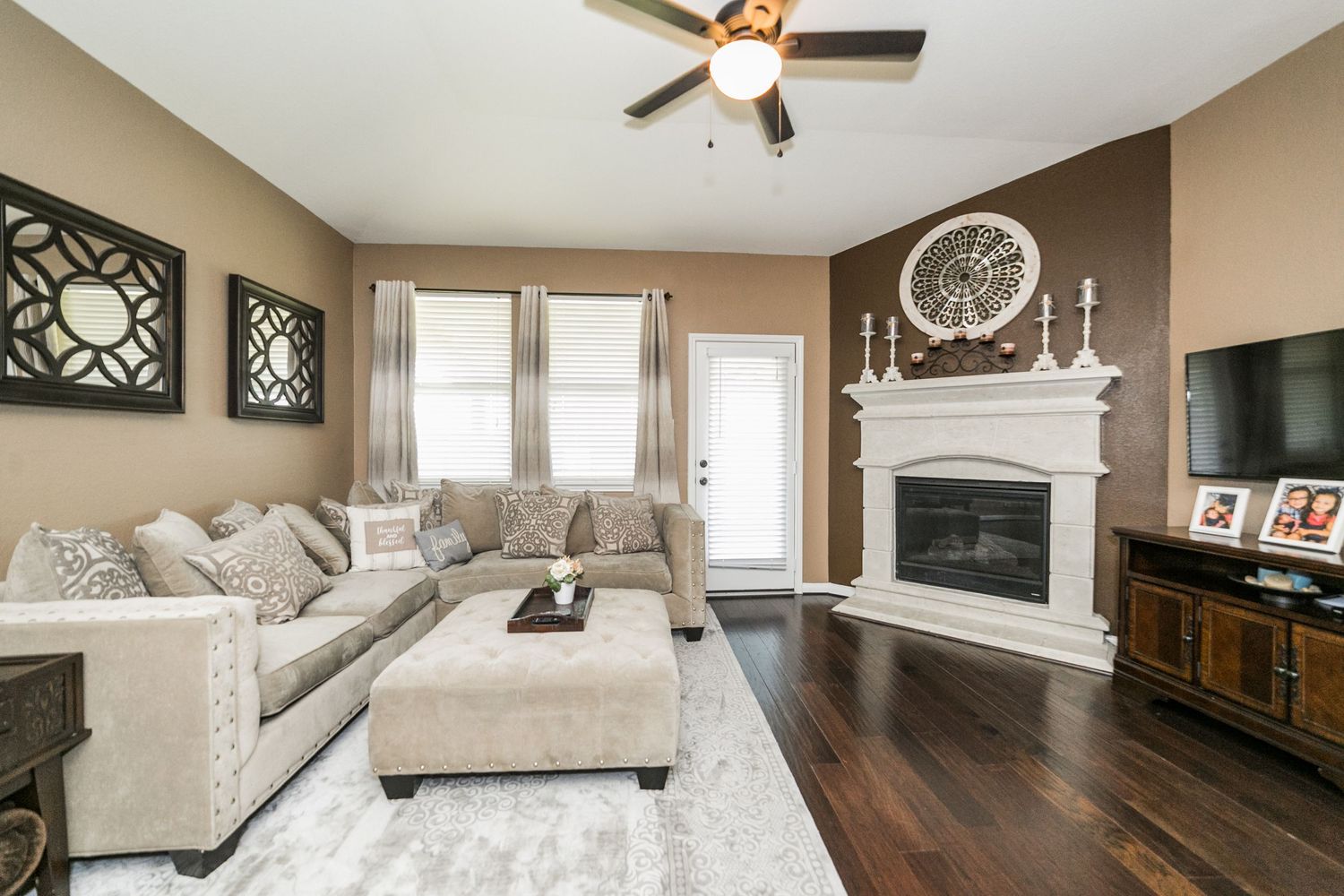 Cozy living room with sectional sofa, white fireplace, hardwood floors, ceiling fan, and large TV on dark wood entertainment center.