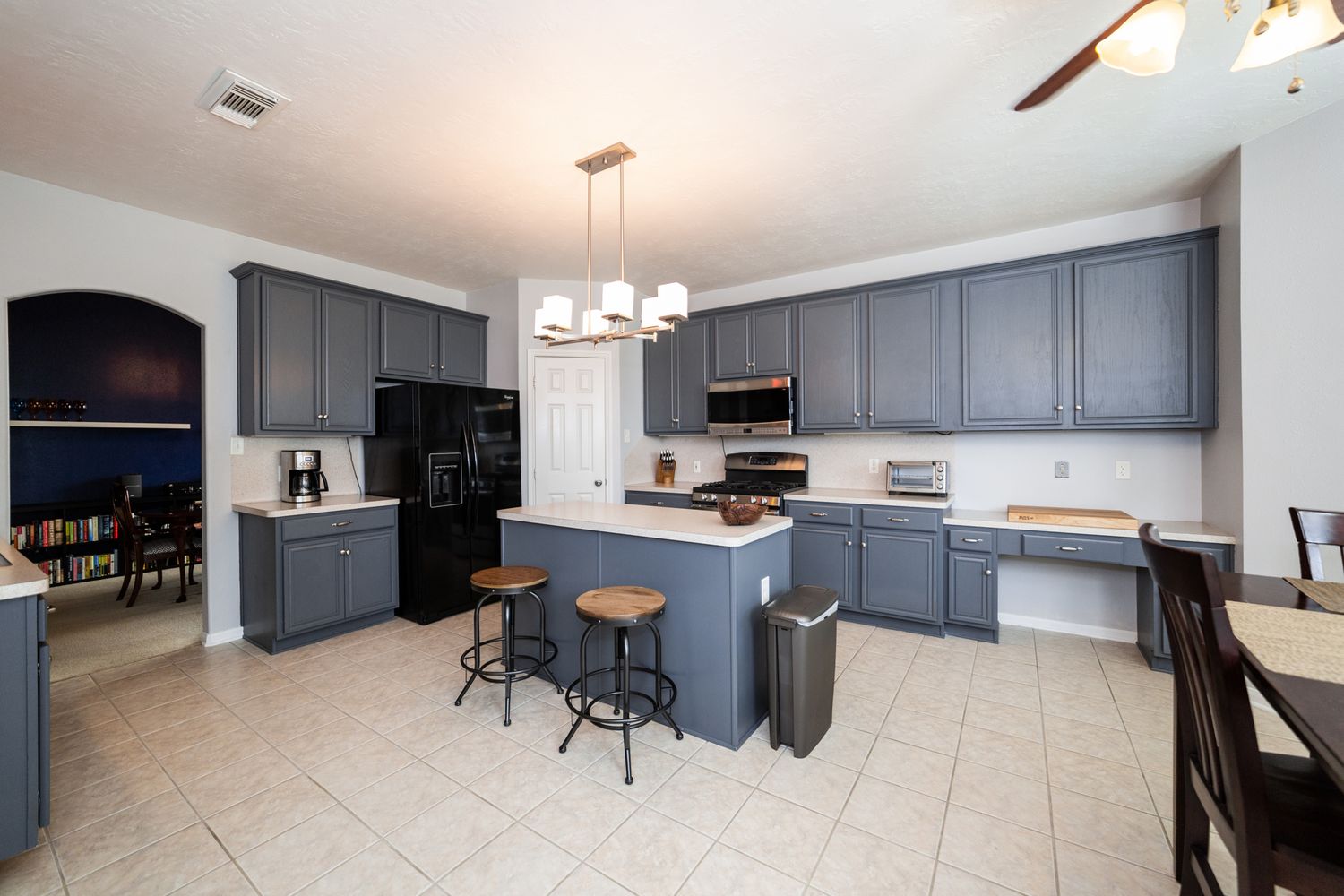 Modern kitchen with gray cabinets, black appliances, island with bar stools, and light tile flooring.
