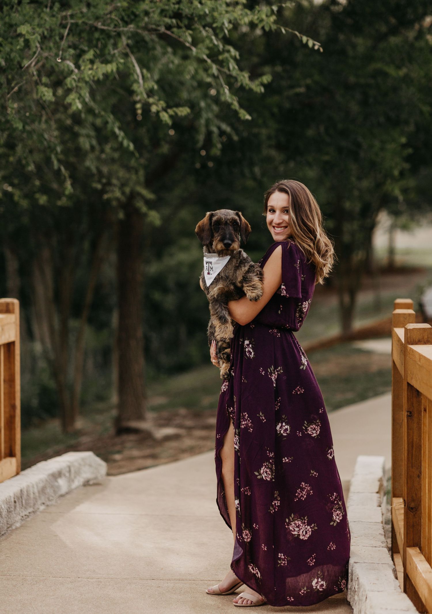 Woman in purple floral maxi dress holds small dog on wooden bridge surrounded by lush green trees.