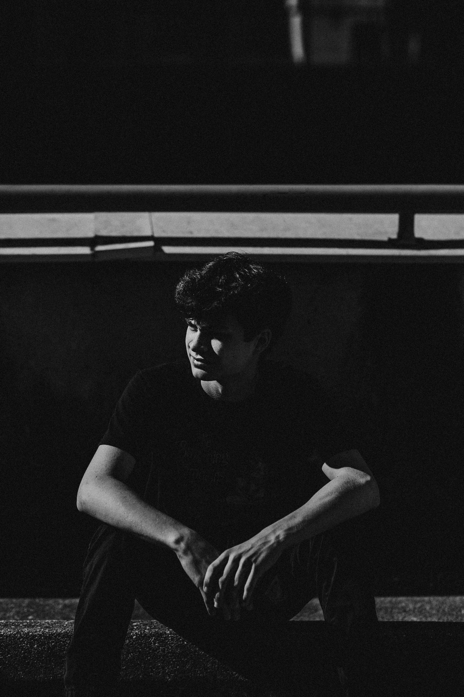 Young man in black t-shirt sitting against a dark wall in dramatic black and white portrait photography.