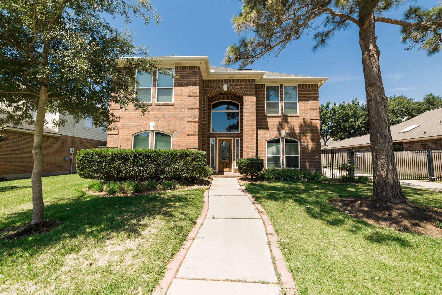 Two-story brick home with manicured lawn, tall trees, and a concrete walkway leading to an elegant arched entrance.