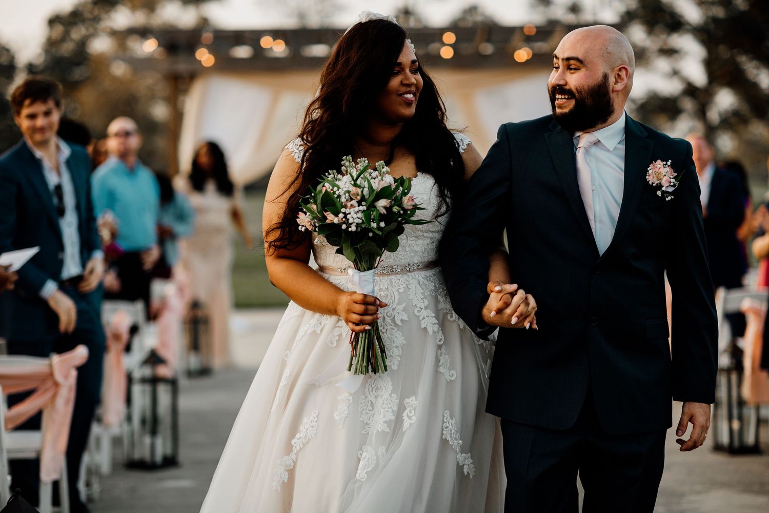 Joyful newlyweds walk down the aisle after wedding ceremony, bride holding floral bouquet in elegant lace gown.