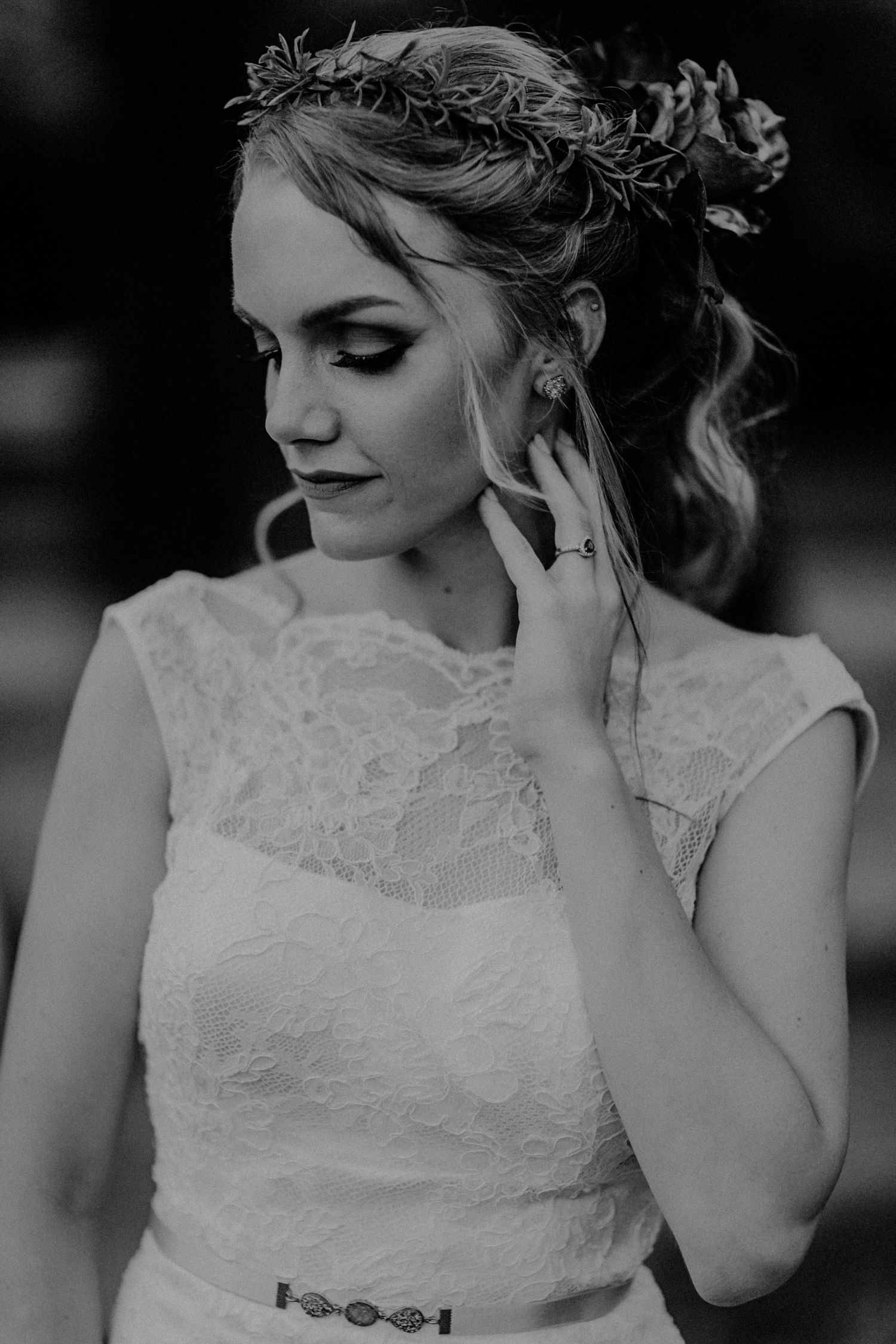 Black and white portrait of a bride in lace dress wearing floral crown, touching her ear, looking down gracefully.