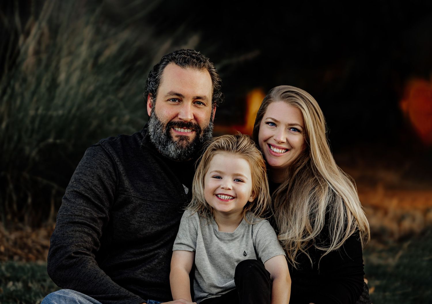 Smiling family of three sitting together outdoors in warm evening light.