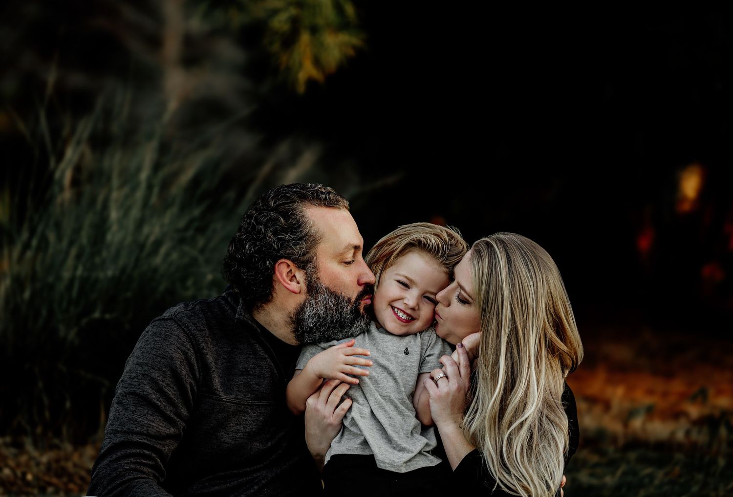 Parents kissing their smiling toddler on the cheeks during a warm family portrait session outdoors.