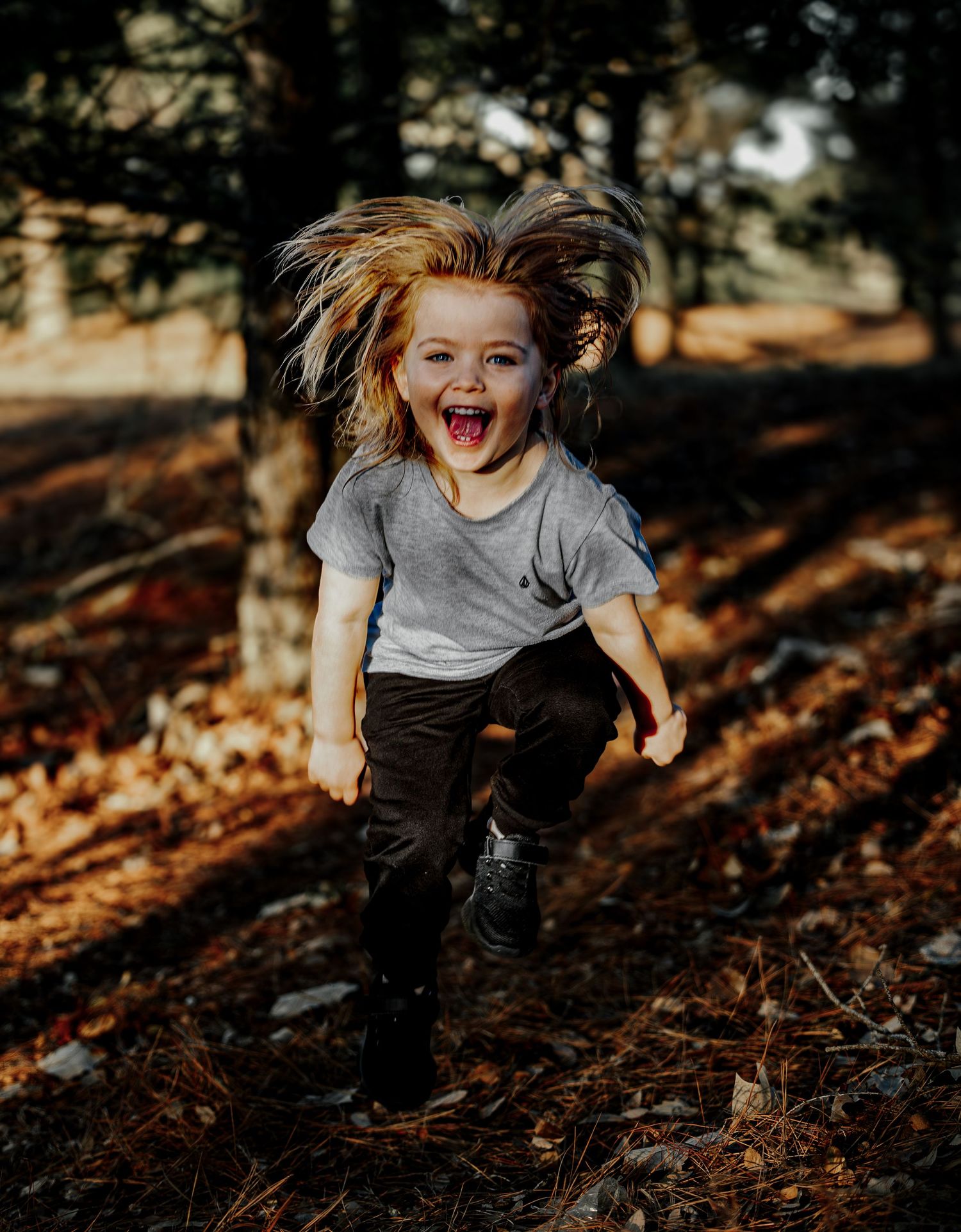 Excited child running joyfully through a forest with hair flying, wearing gray shirt and dark pants.