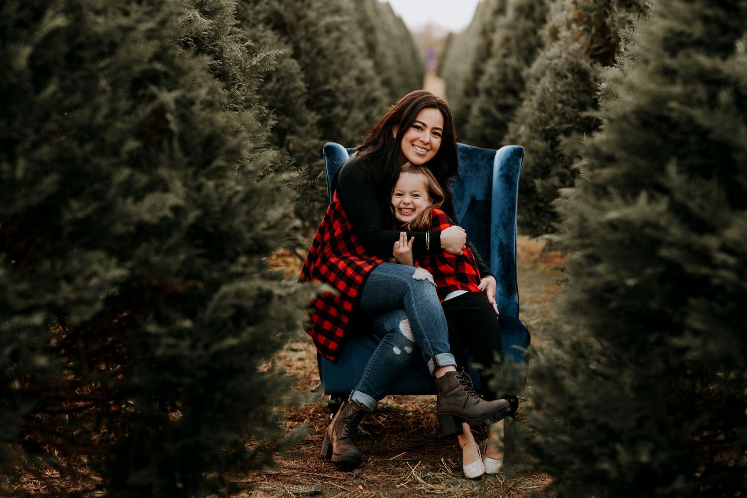 Mother and child sitting in blue velvet chair at Christmas tree farm, wearing red plaid outfits, smiling.