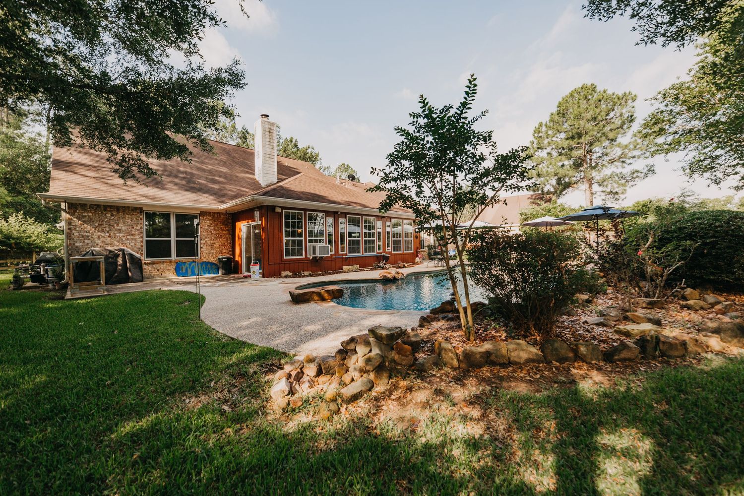 Brick home with a backyard pool, patio, and lush green lawn surrounded by tall trees on a sunny day.