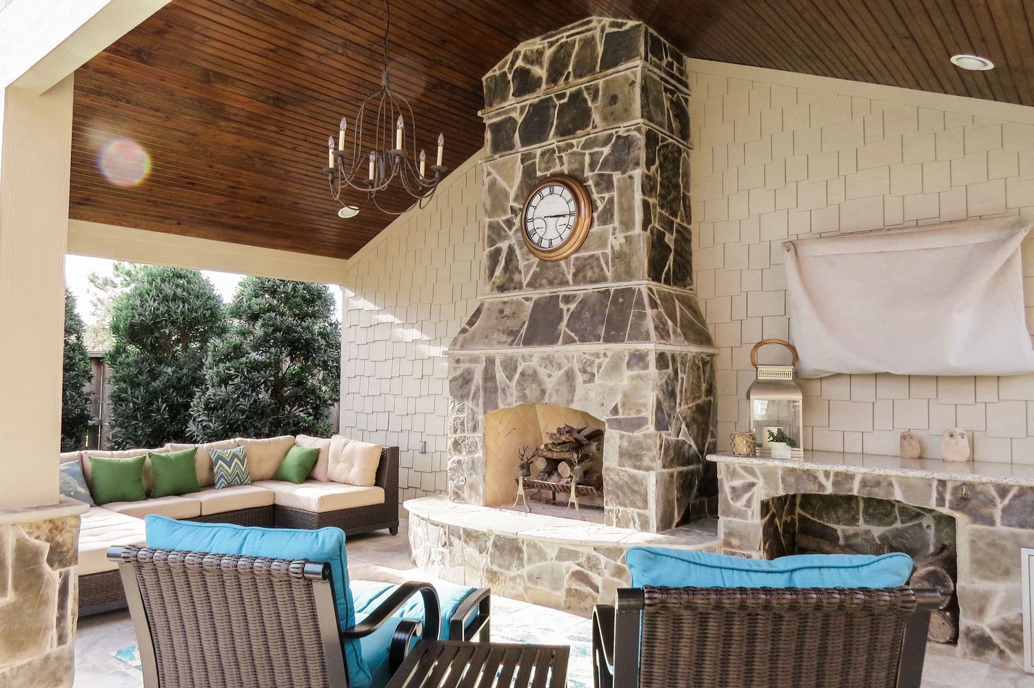 Covered outdoor patio with stone fireplace, teal cushioned seating, wood ceiling, and chandelier.