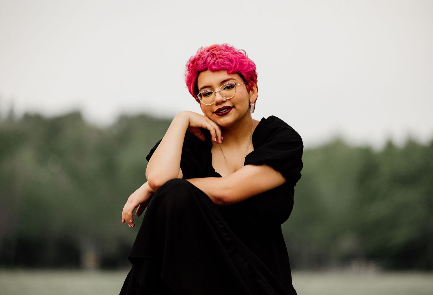 Woman with vibrant pink short hair wearing black outfit, posing confidently outdoors with green trees in background.
