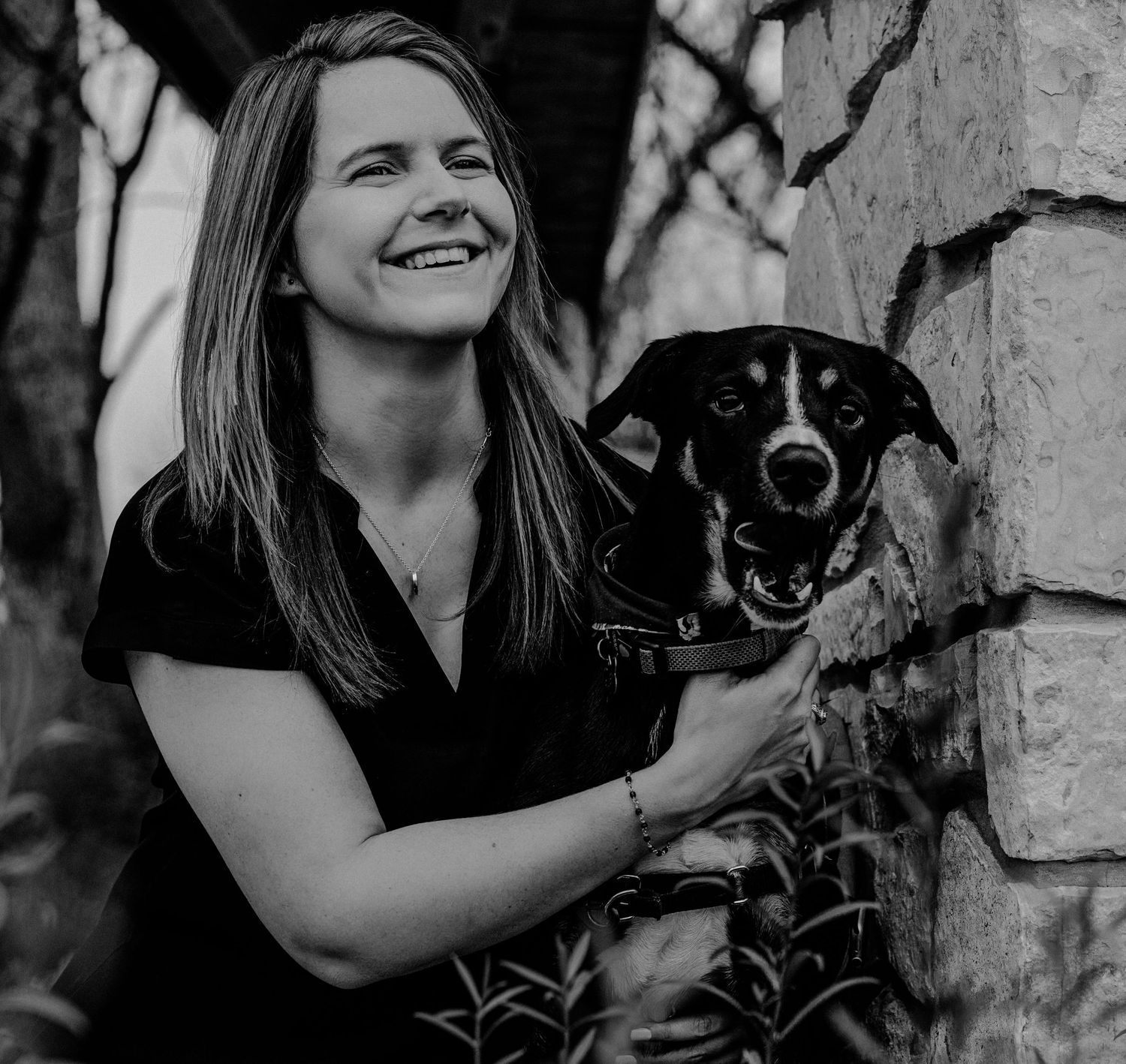 Black and white photo of a smiling woman posing outdoors with her black and white dog near a stone wall.