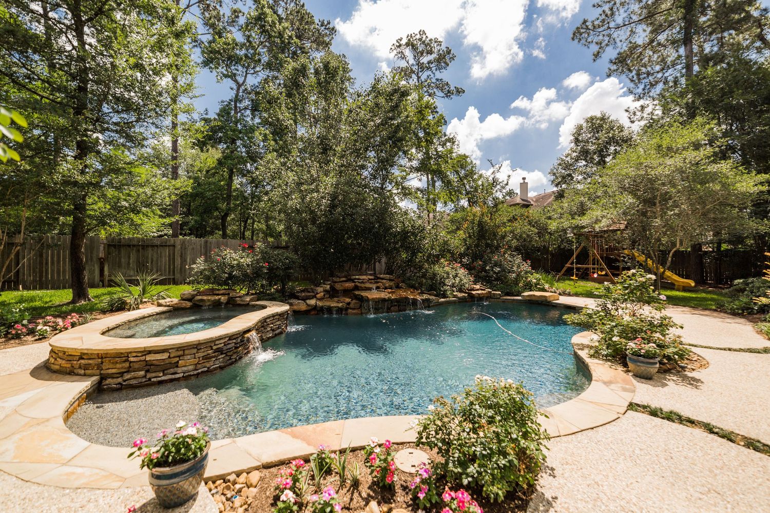 Luxurious freeform backyard pool with spa, stone decking, lush landscaping, and mature trees in a private setting.