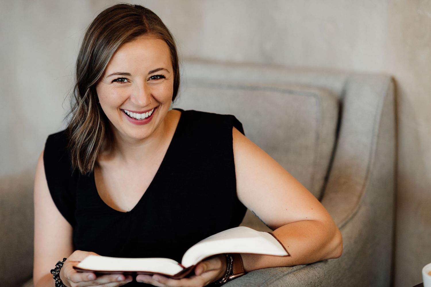 Smiling woman in black top sitting in gray chair holding open book, bright natural light background.
