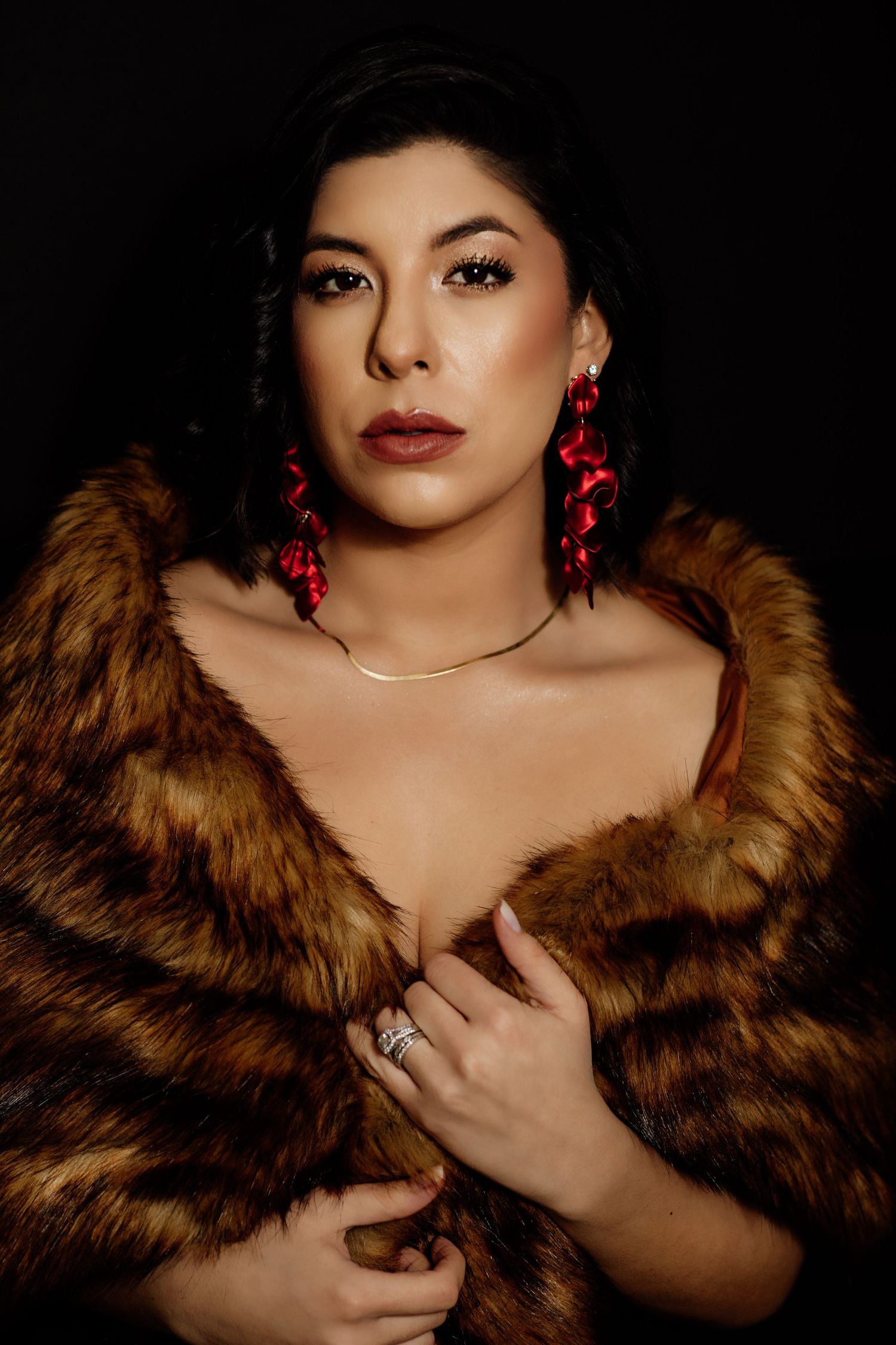 Elegant woman in fur coat with red earrings posing against a dark background.