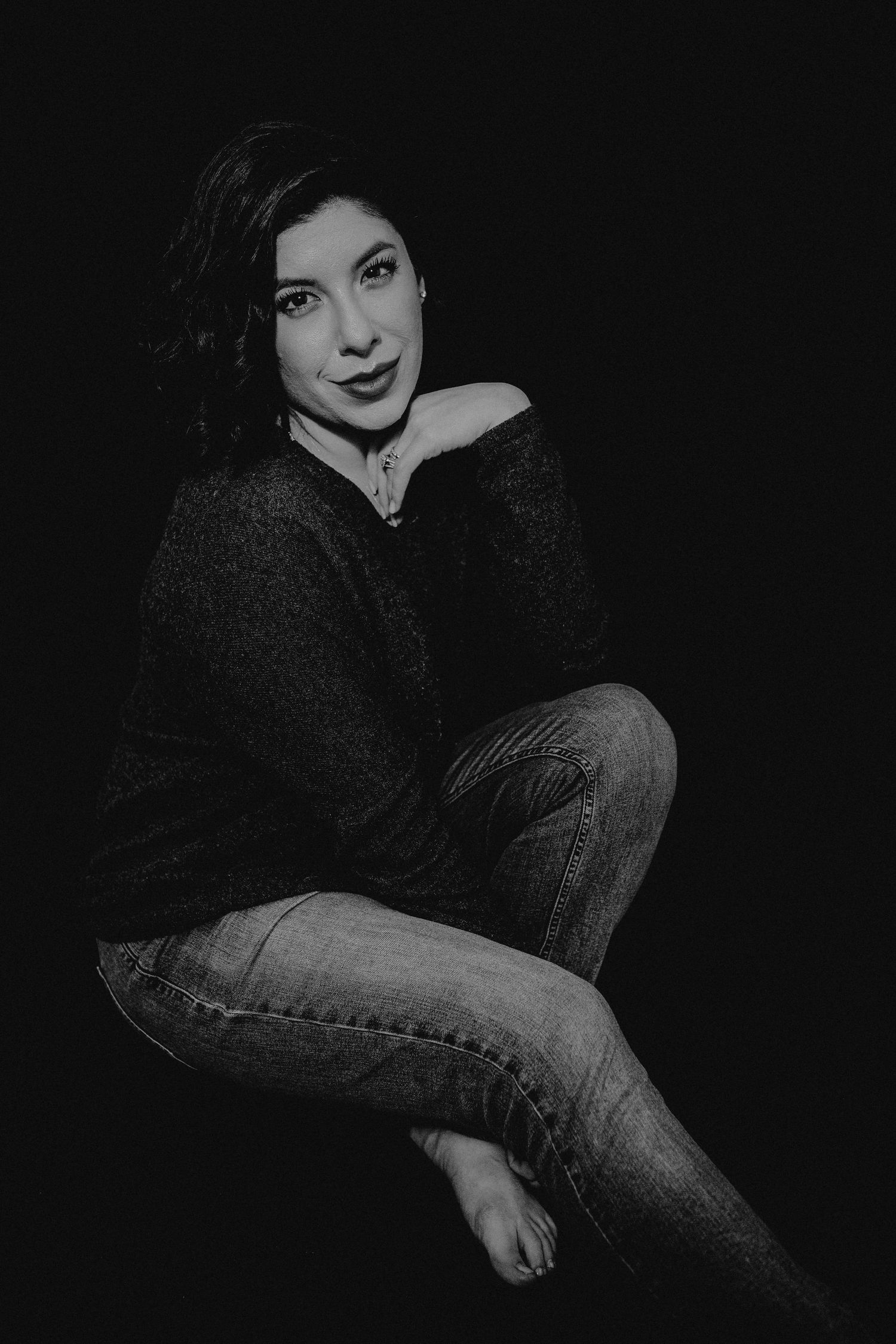 Black and white portrait of a woman in a sweater and jeans sitting gracefully against a dark background.