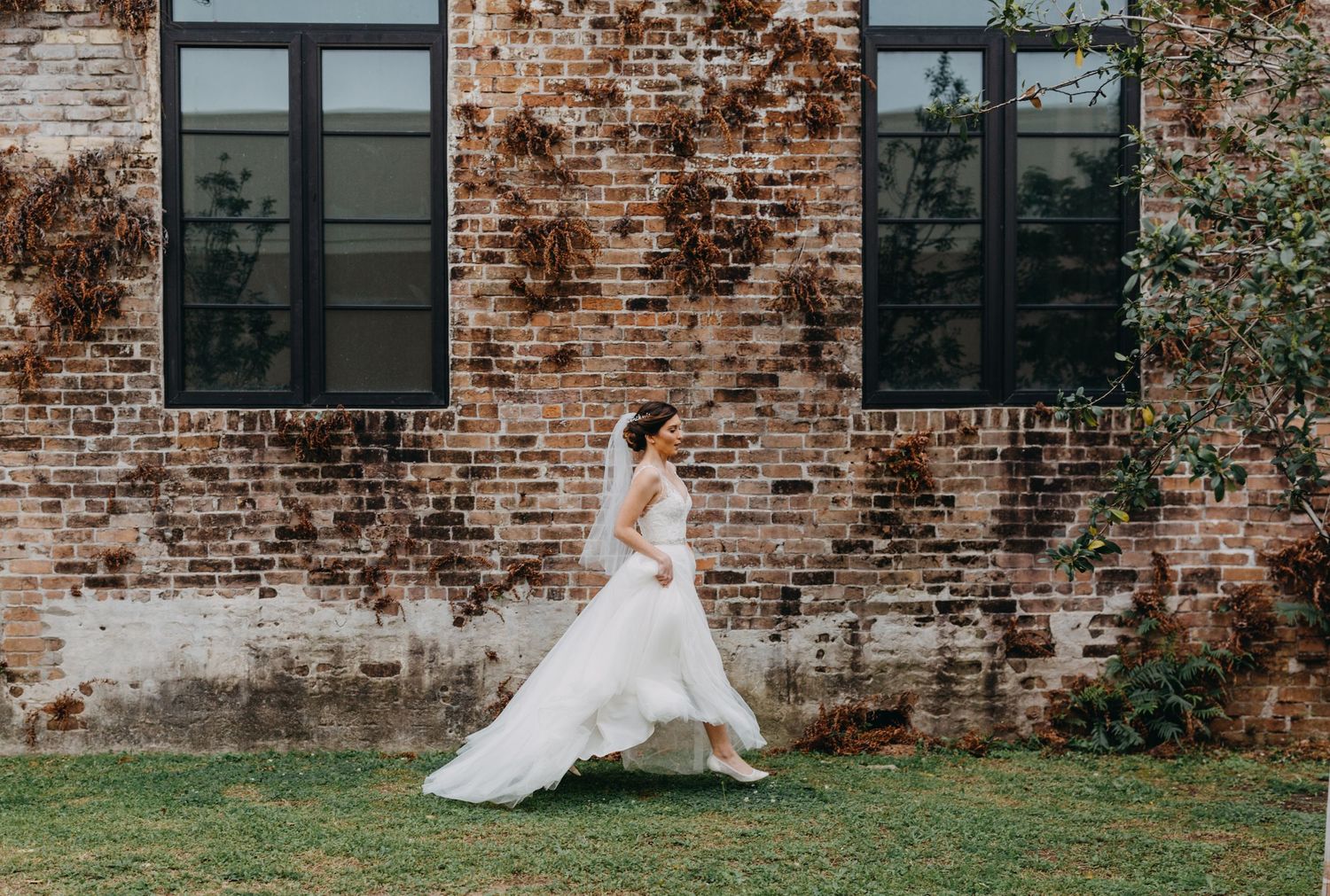 Bride in a flowing white wedding gown and veil walking gracefully beside a rustic brick building.