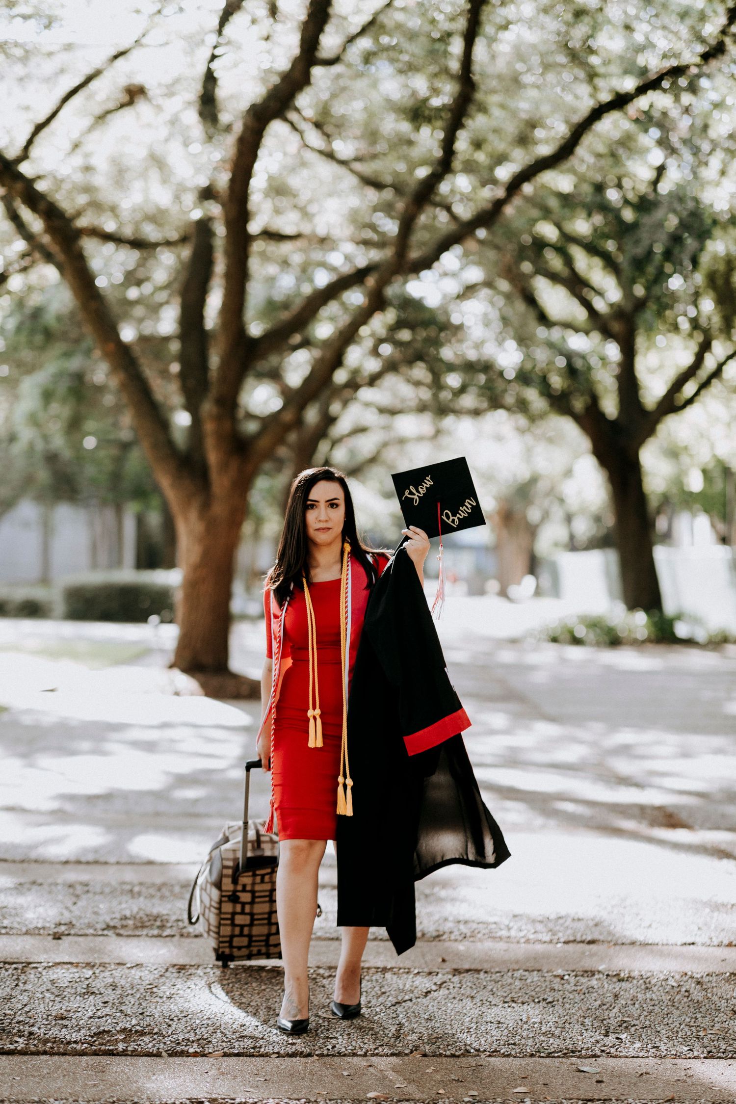 A young woman in a red dress and black graduation gown holds her cap aloft outdoors among tree-lined paths.