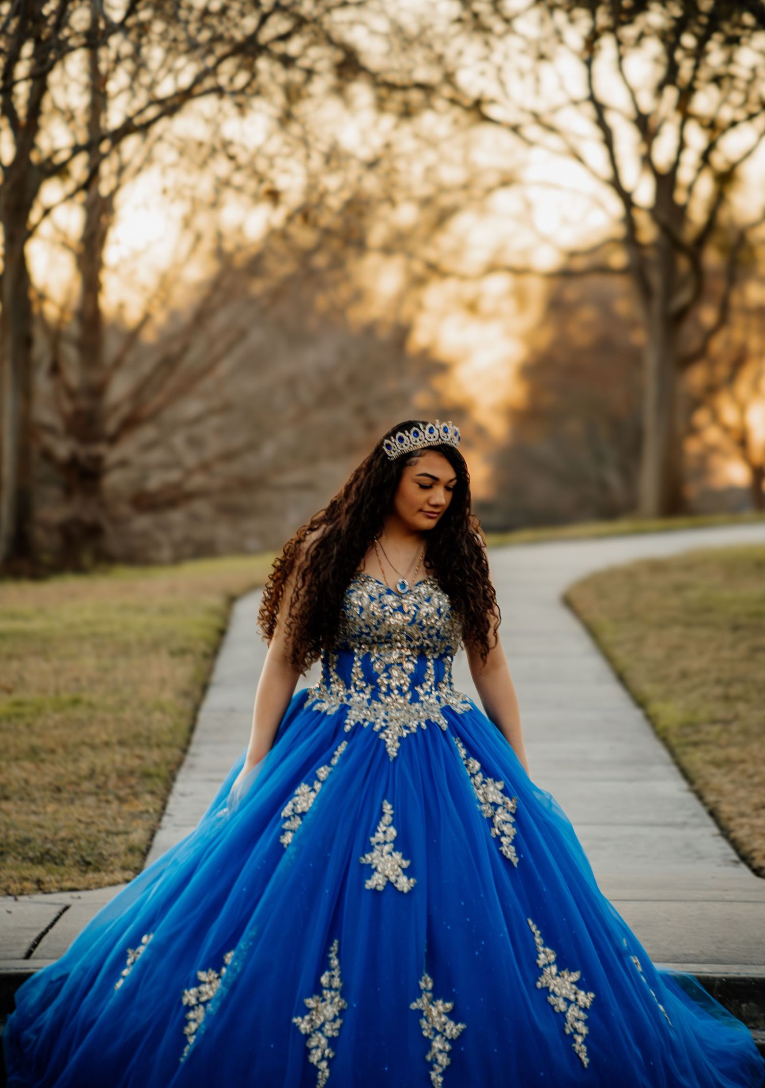 A young woman in a royal blue quinceañera ball gown and tiara poses on a garden path.