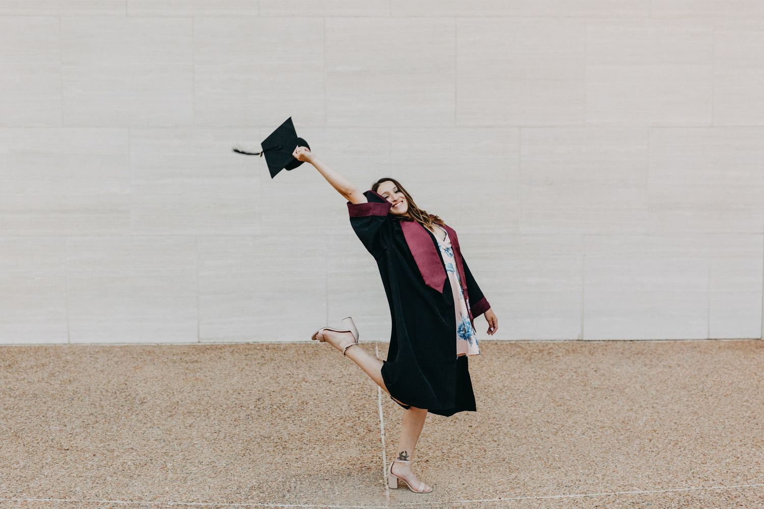 A joyful graduate in black gown and pink sash tosses cap in the air outdoors.