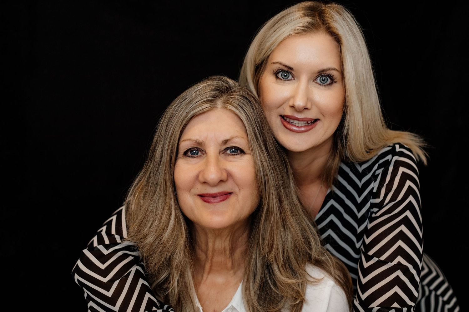 Two blonde women smiling together in black and white tops on dark background.