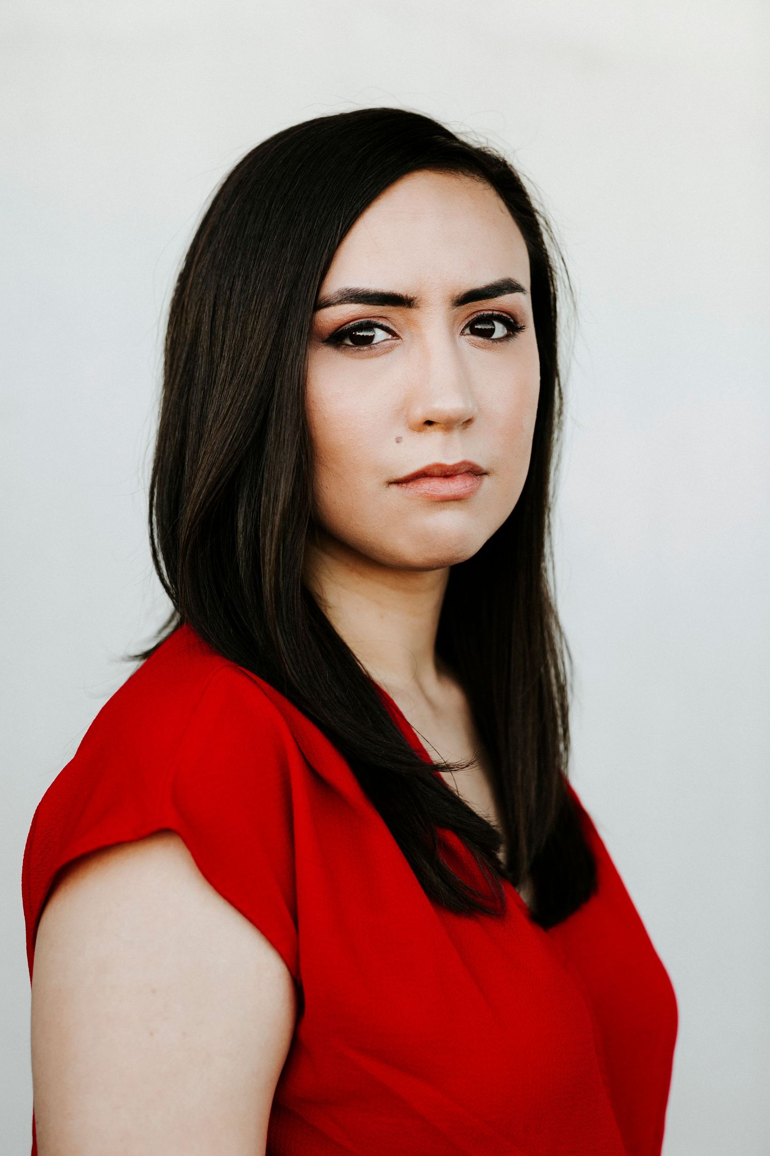 Young woman with long dark hair wearing a red top poses against a light gray background.