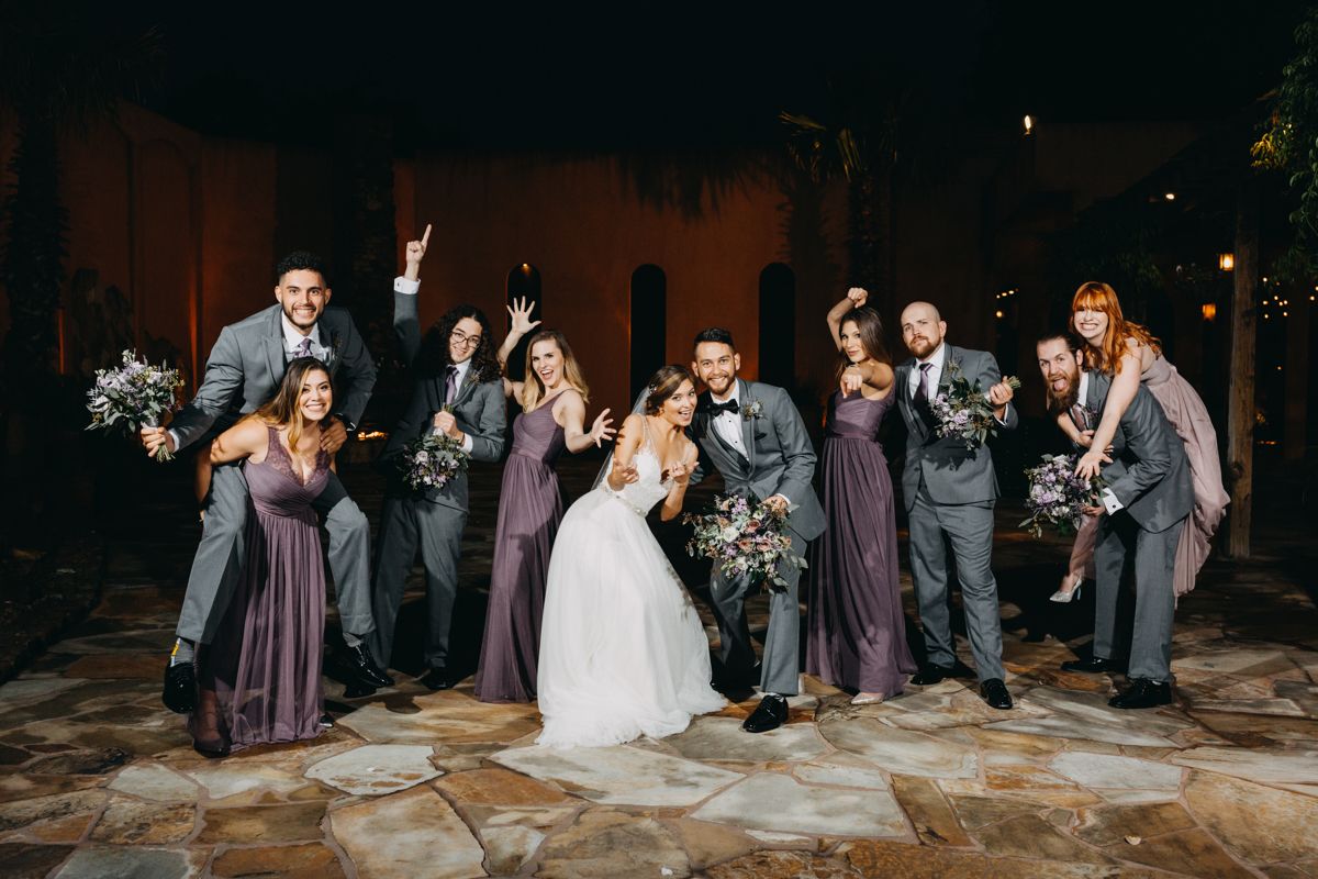 Wedding party celebrating outdoors at night, bridesmaids in purple gowns and groomsmen in gray suits with bride and groom.