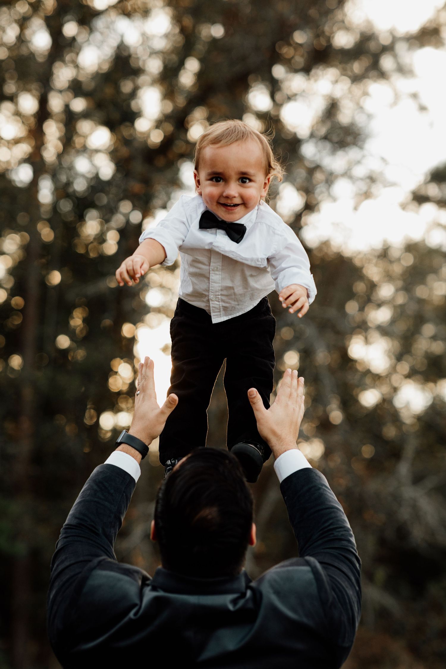 Joyful baby in bow tie tuxedo being lifted by father outdoors in golden light.