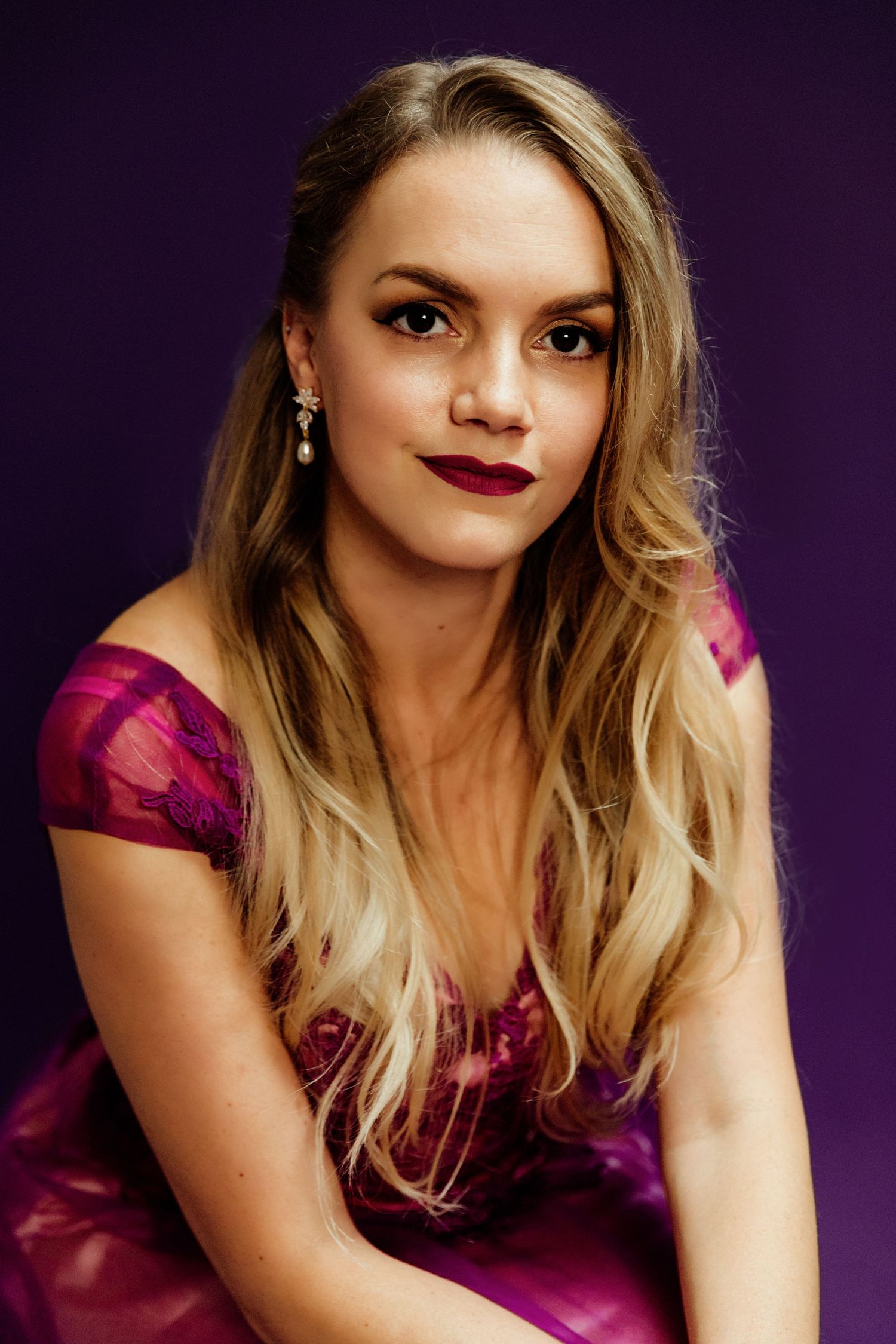 Young blonde woman in magenta off-shoulder gown with dark lipstick poses against purple studio backdrop.
