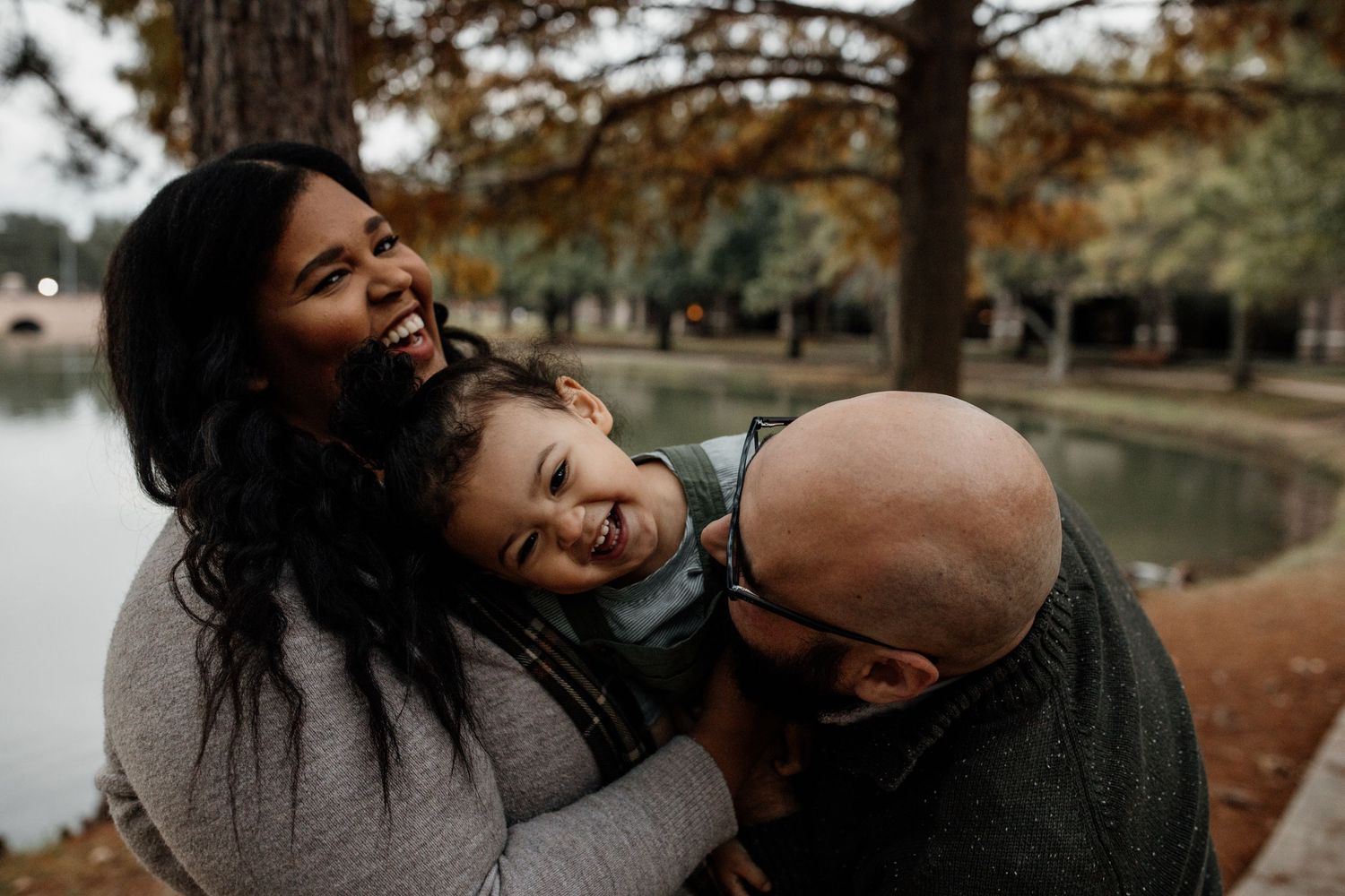 A joyful family of three laughing together outdoors near a pond surrounded by autumn trees.