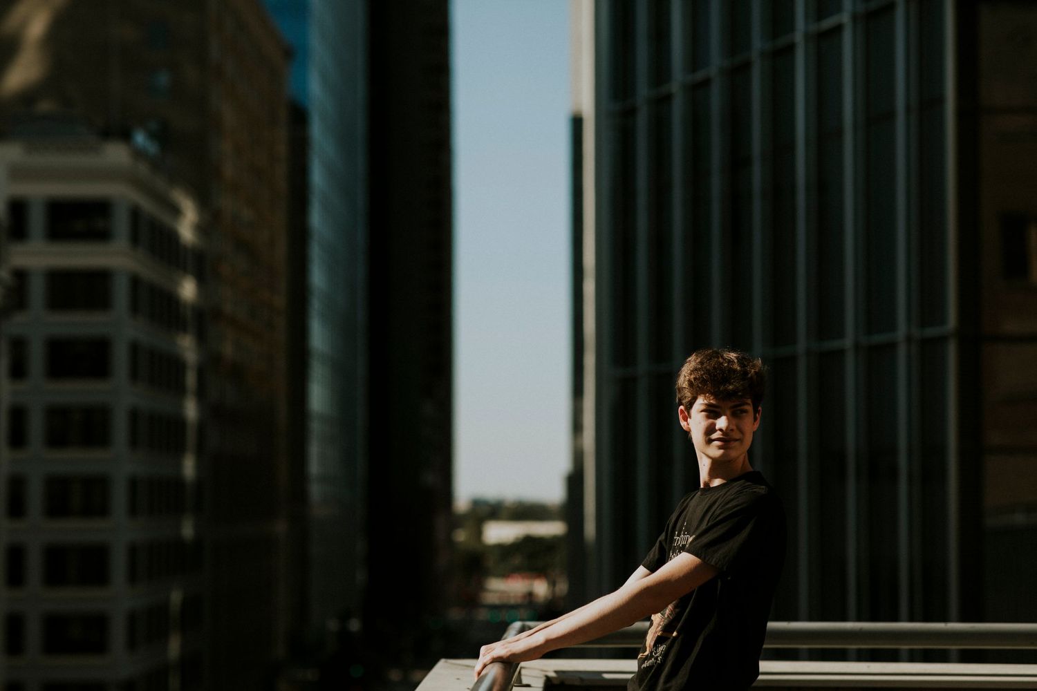 Young man in black t-shirt smiling on urban rooftop with modern glass skyscrapers in background.