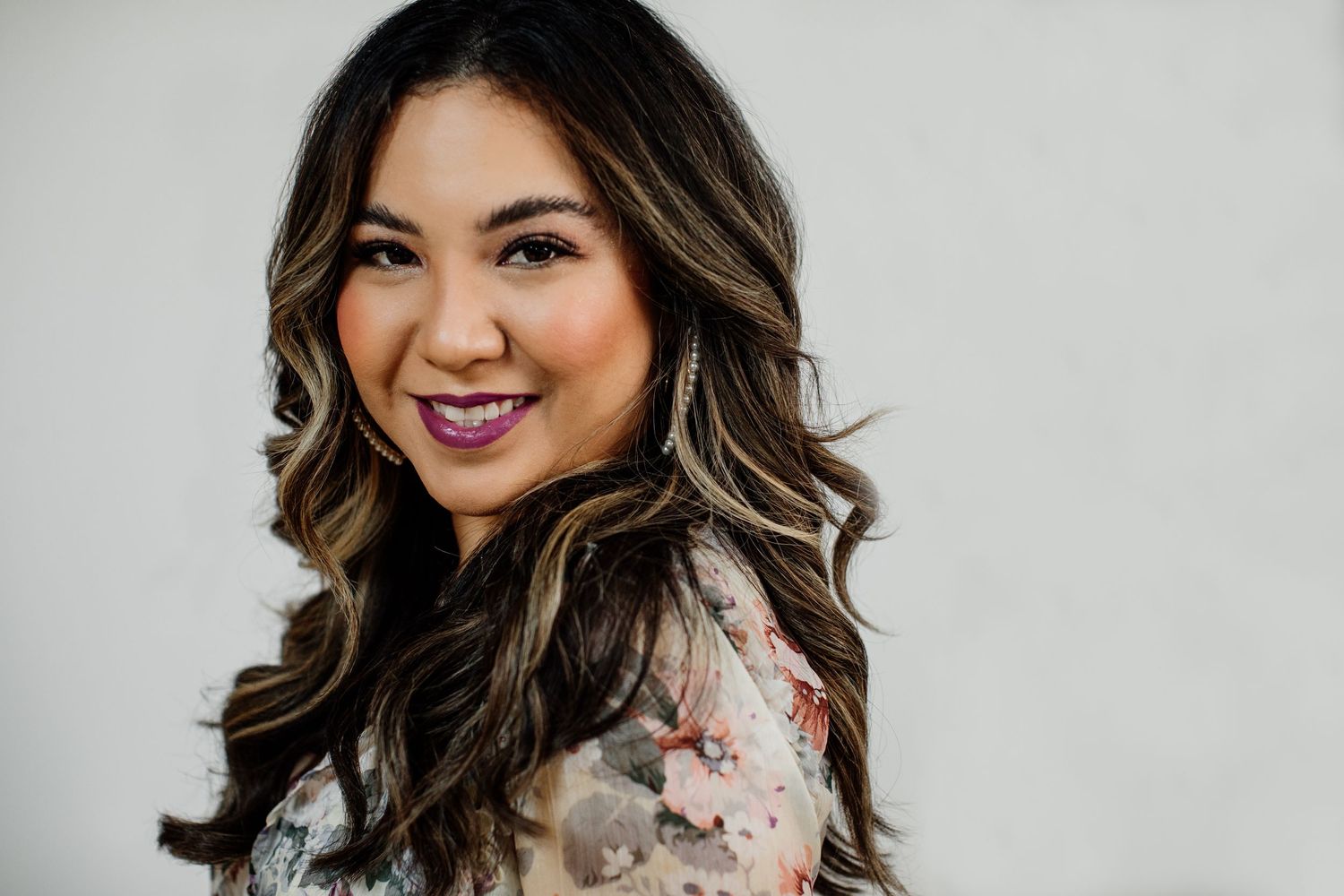 Smiling young woman with highlighted wavy hair wearing a floral top, posing against a light gray background.