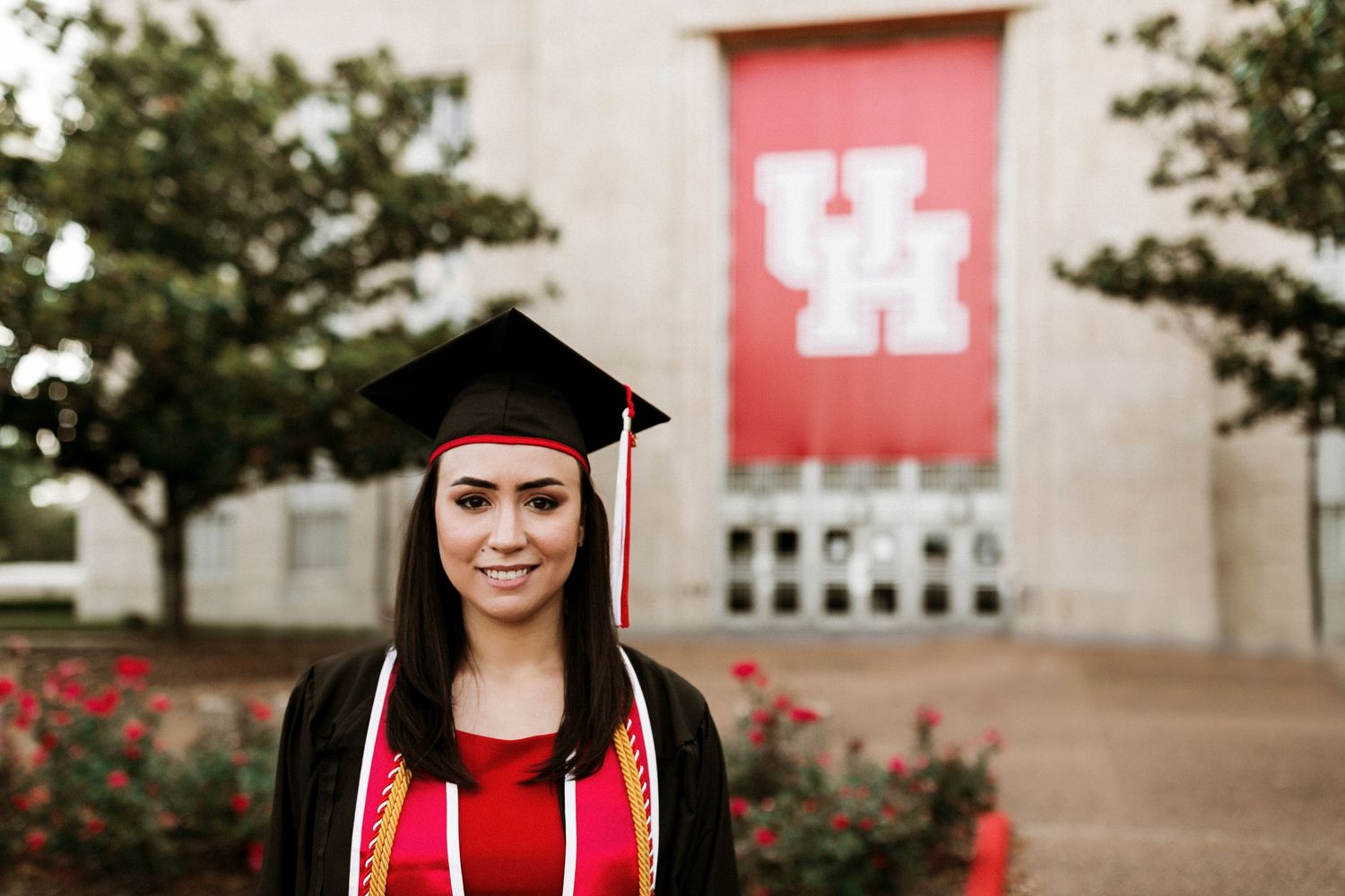 Graduate in cap and gown smiles before University of Houston red UH banner at commencement ceremony.