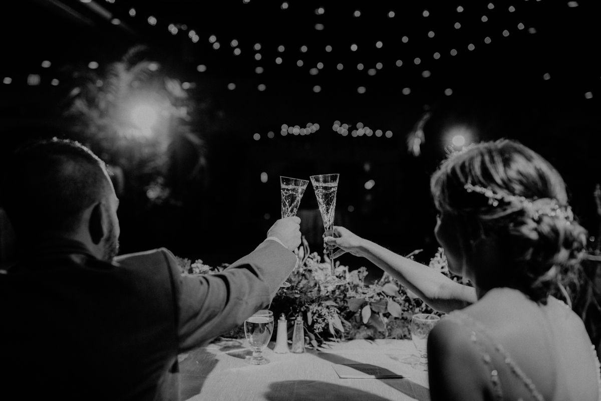 Bride and groom toasting champagne glasses at wedding reception under string lights, black and white.
