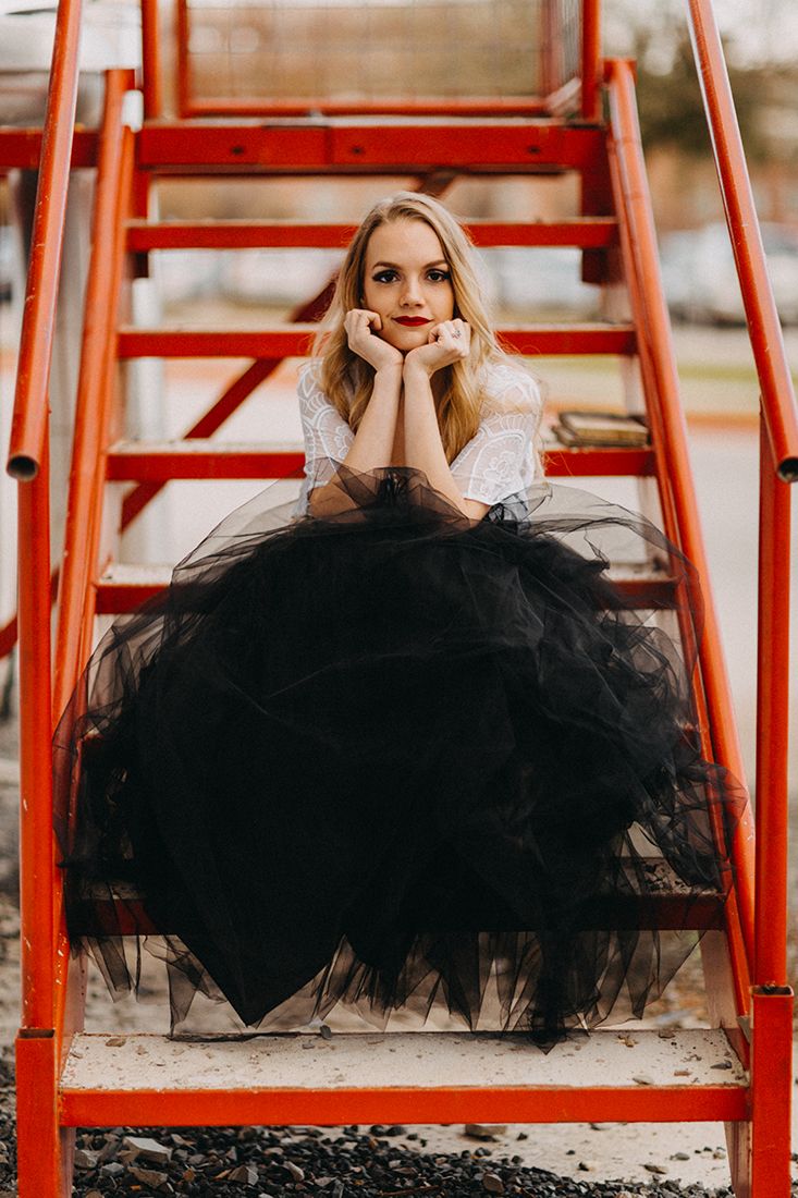 Young blonde woman in white top and black tulle skirt sitting gracefully on red metal stairs outdoors.