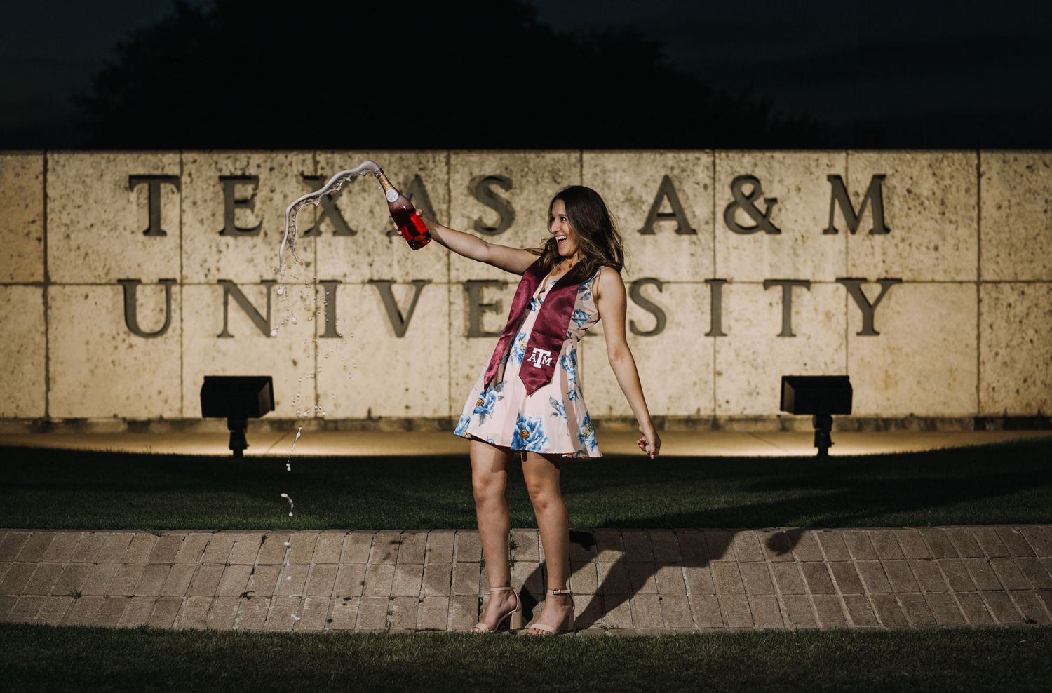 Young woman in floral dress posing in front of Texas A&M University sign at night.