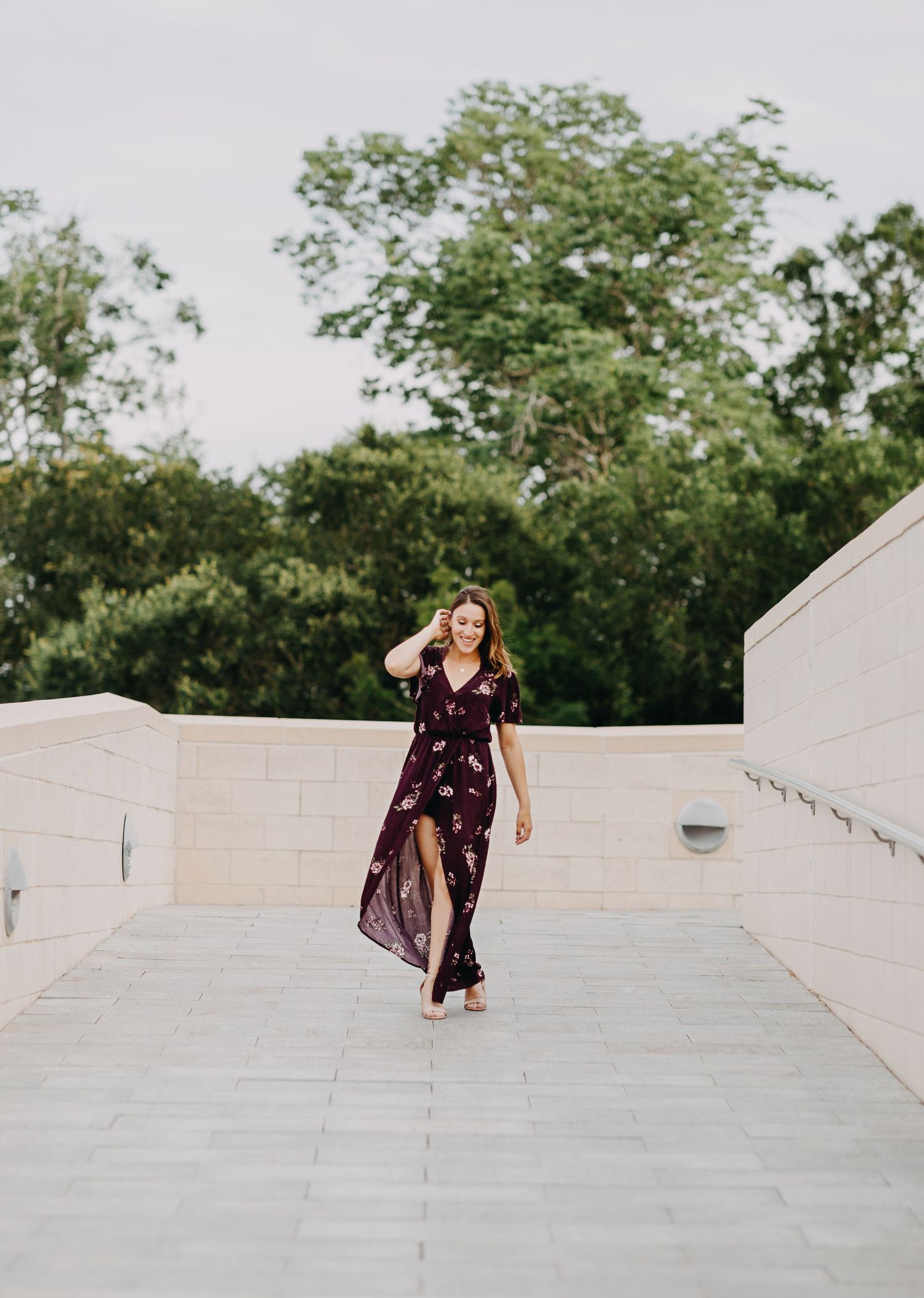 A young woman in a floral burgundy maxi dress walks gracefully on a stone terrace with lush green trees behind her.