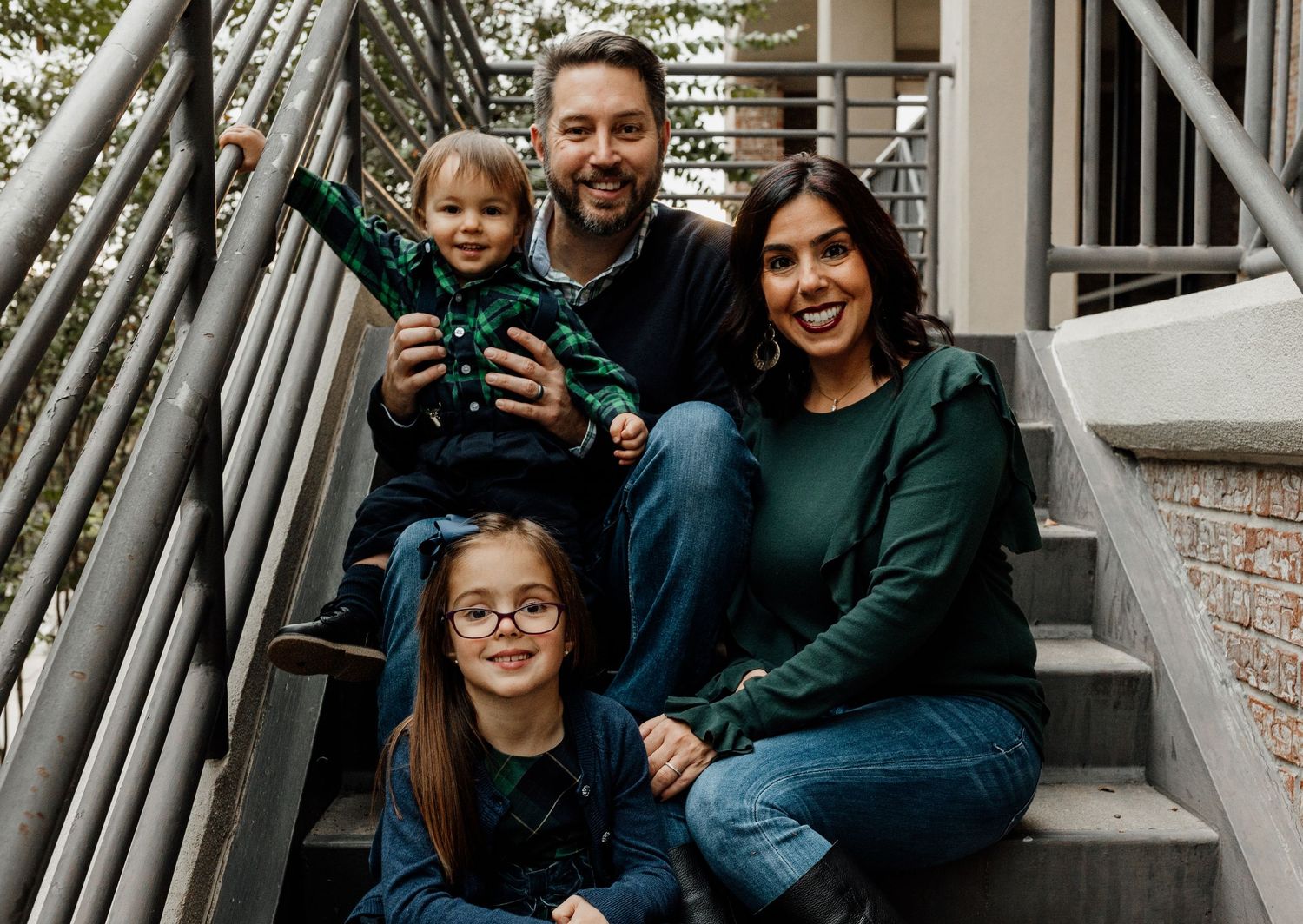 Family of four in matching green outfits smiling on outdoor steps, parents with two young daughters in a warm portrait.