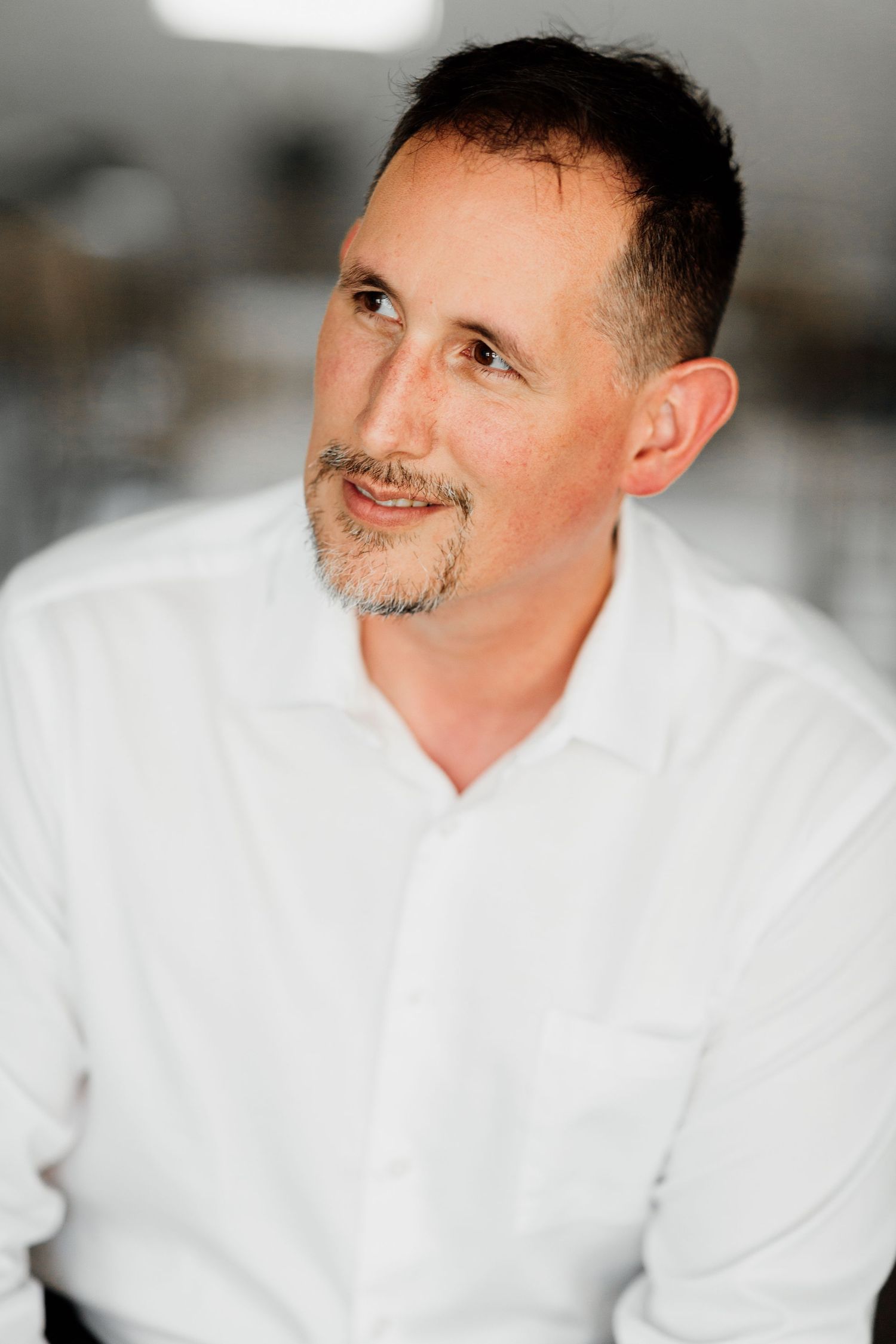 Middle-aged man with short hair and goatee wearing a white button-up shirt, smiling softly in a bright indoor setting.