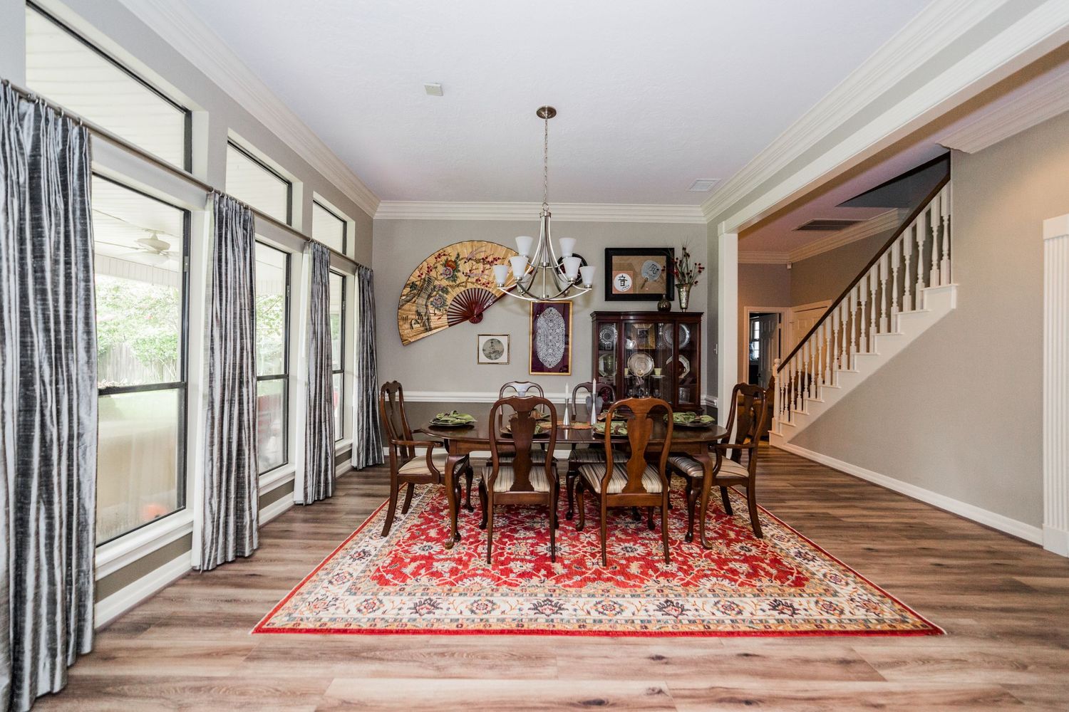Elegant dining room with hardwood floors, red Persian rug, chandelier, large windows, and a wooden staircase.