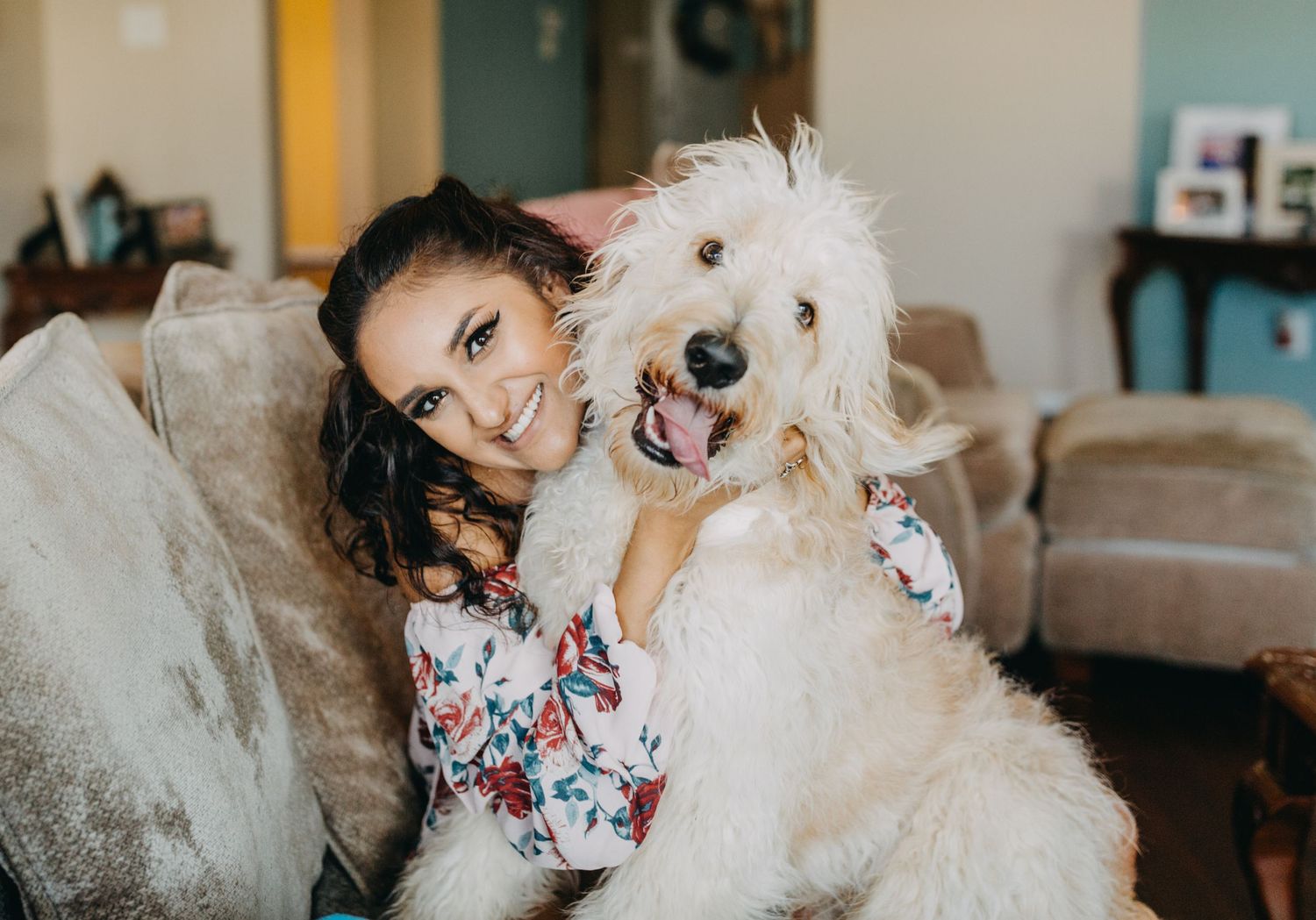 Smiling young woman hugging a fluffy white Goldendoodle dog on a cozy sofa.