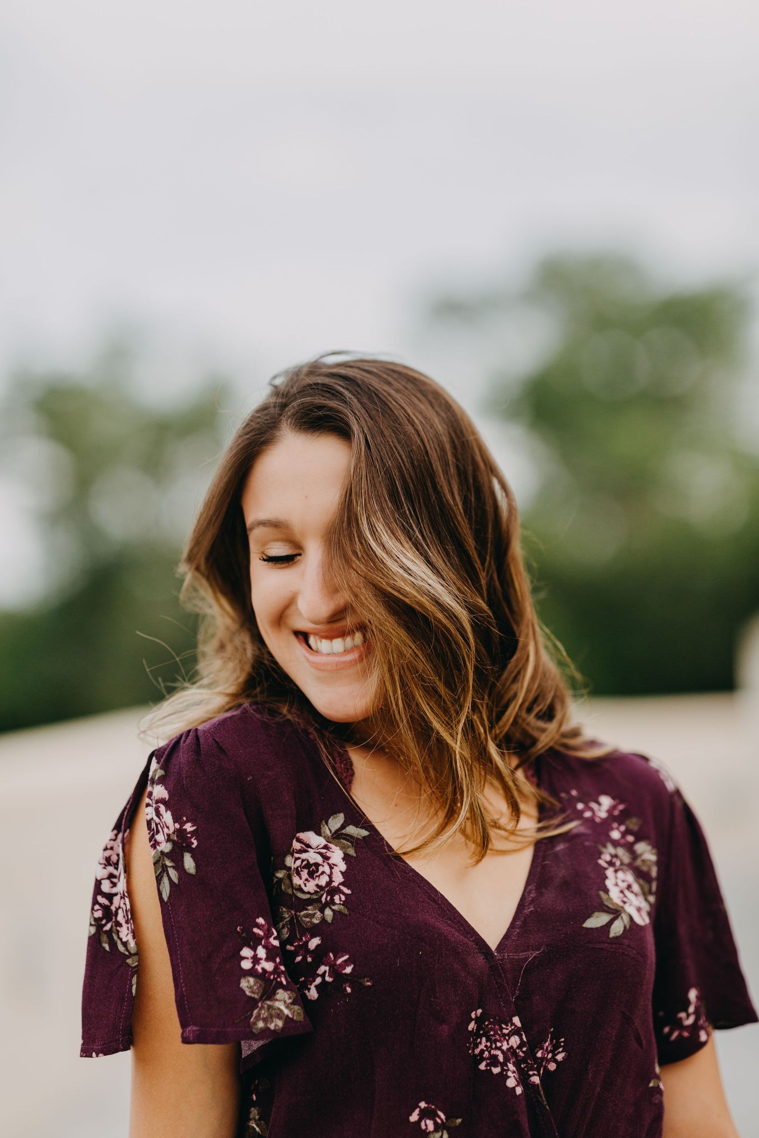Young woman in purple floral dress smiles warmly outdoors with soft bokeh green background.