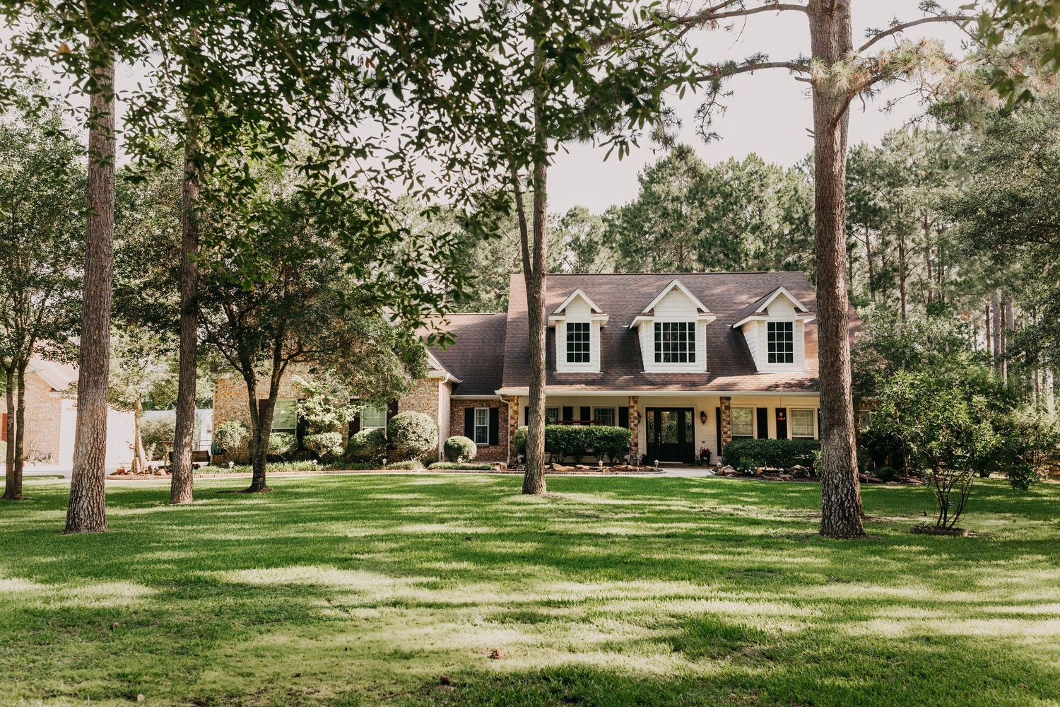 Two-story brick home with white trim surrounded by tall pine trees and lush green lawn in a wooded suburban setting.