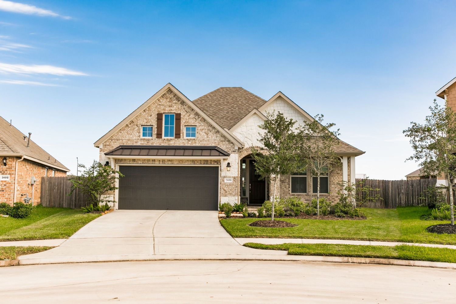 Brick two-story suburban home with dark garage door, manicured lawn, and lush landscaping on a quiet residential street.