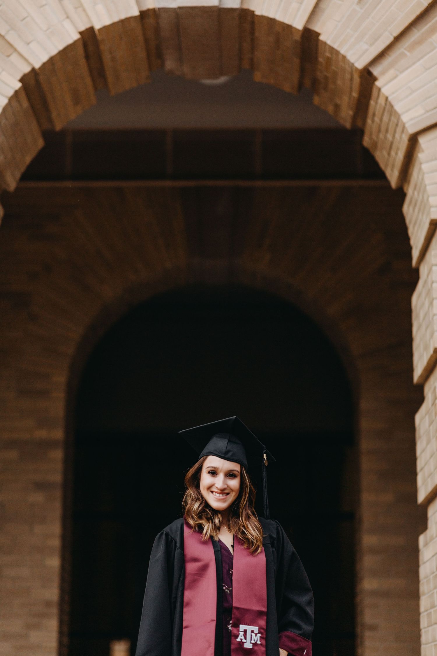 Smiling graduate in cap and gown with Texas A&M logo stands under stone archway for graduation portrait.