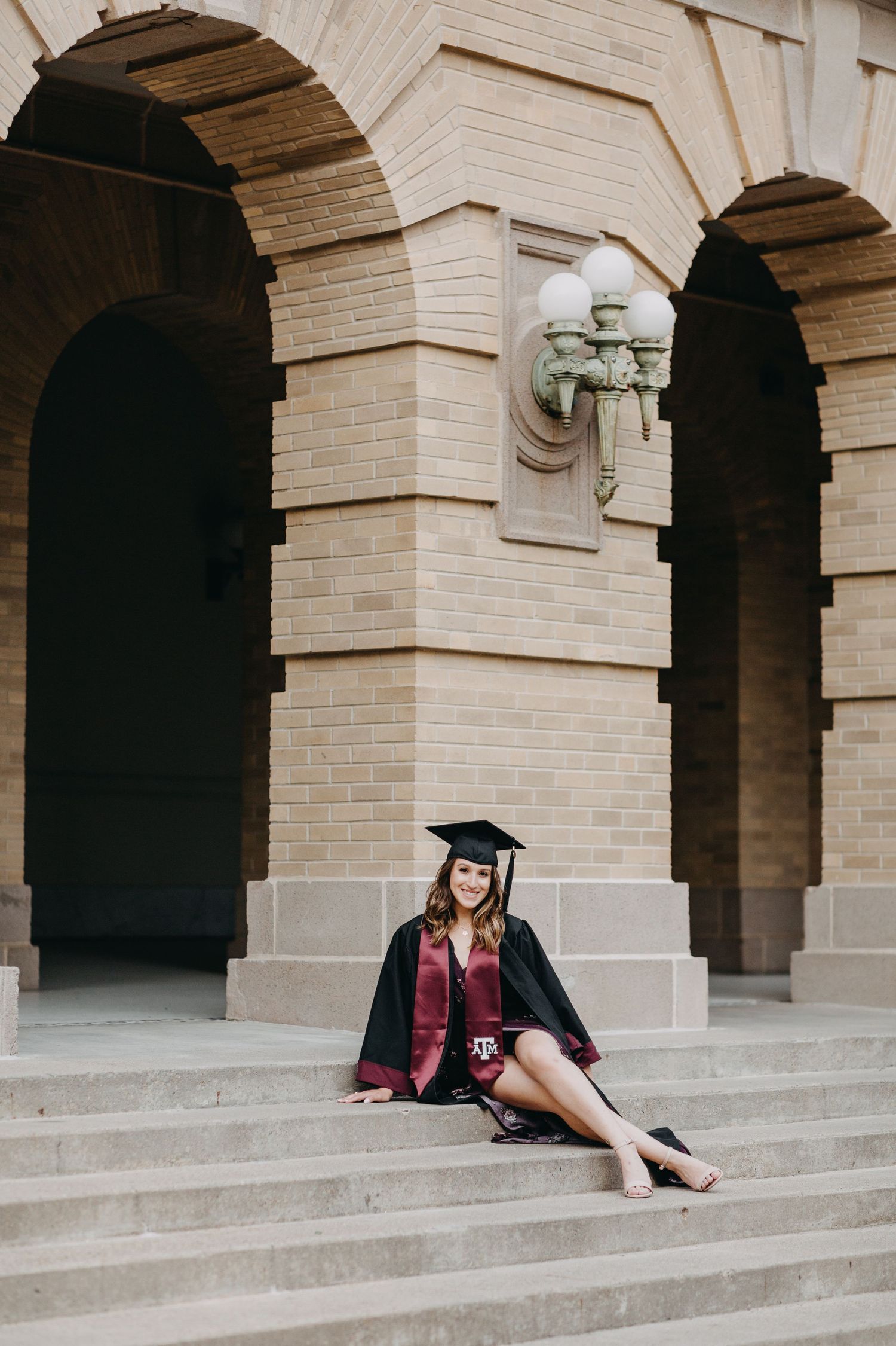 A smiling graduate in black cap and gown with maroon regalia sits on stone steps of a brick university building.